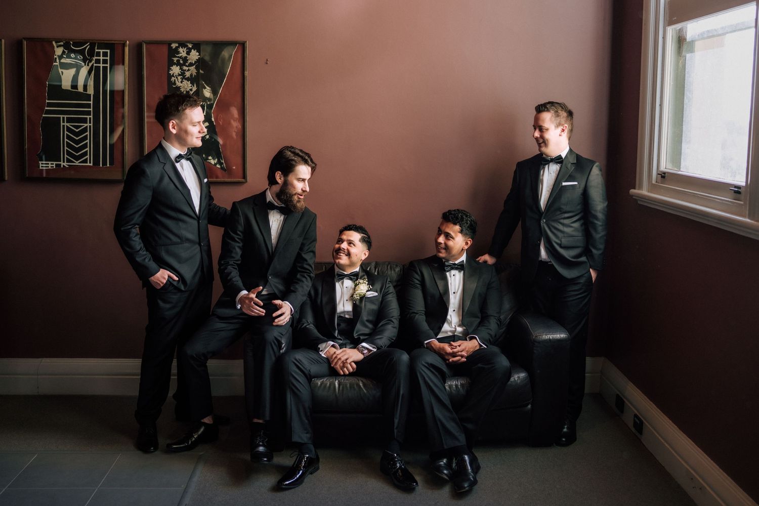 Five groomsmen in black tuxedos pose elegantly in a moody, dimly lit room before a wedding ceremony.