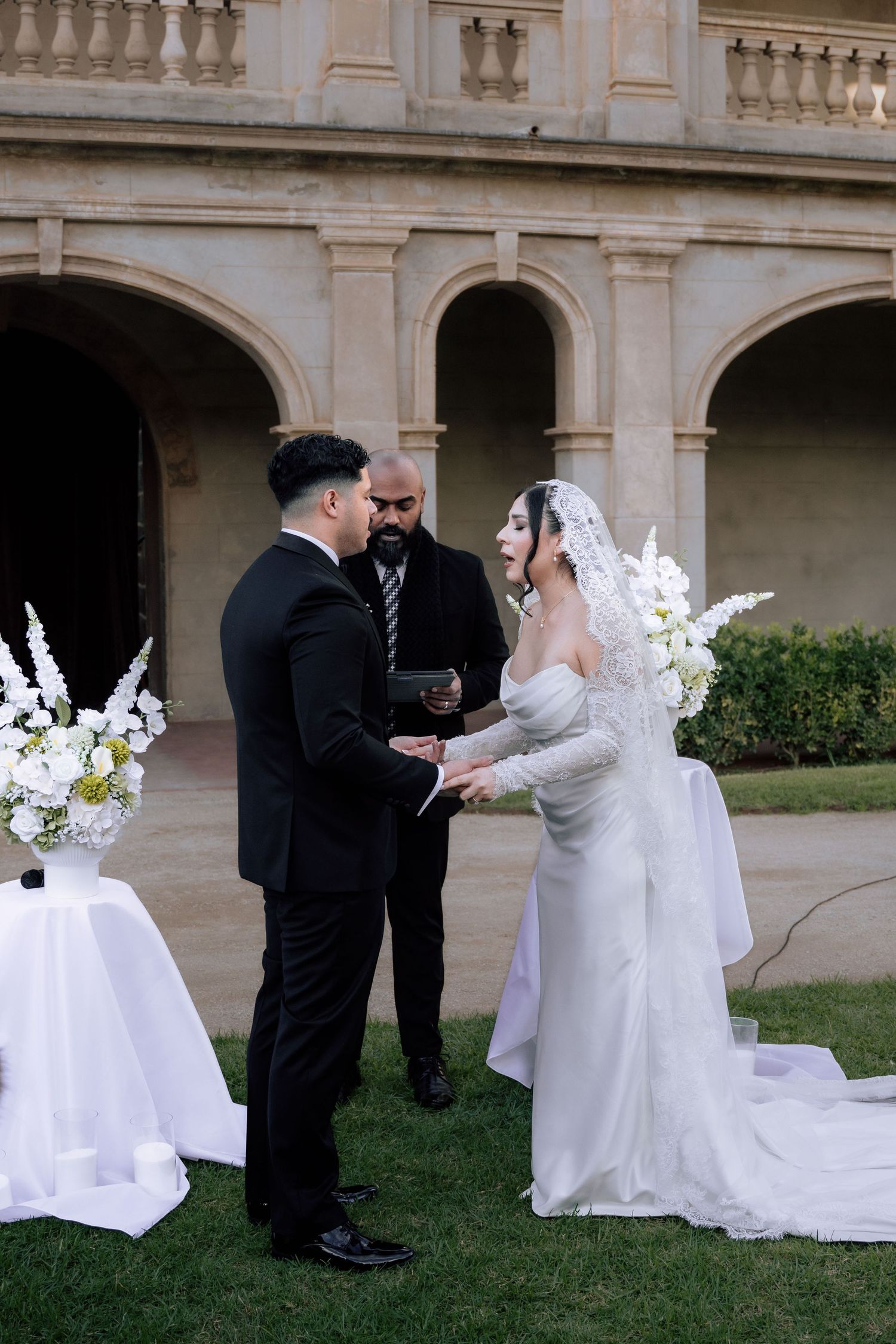 Bride and groom exchange vows outdoors at elegant venue with arched colonnade and white floral arrangements.