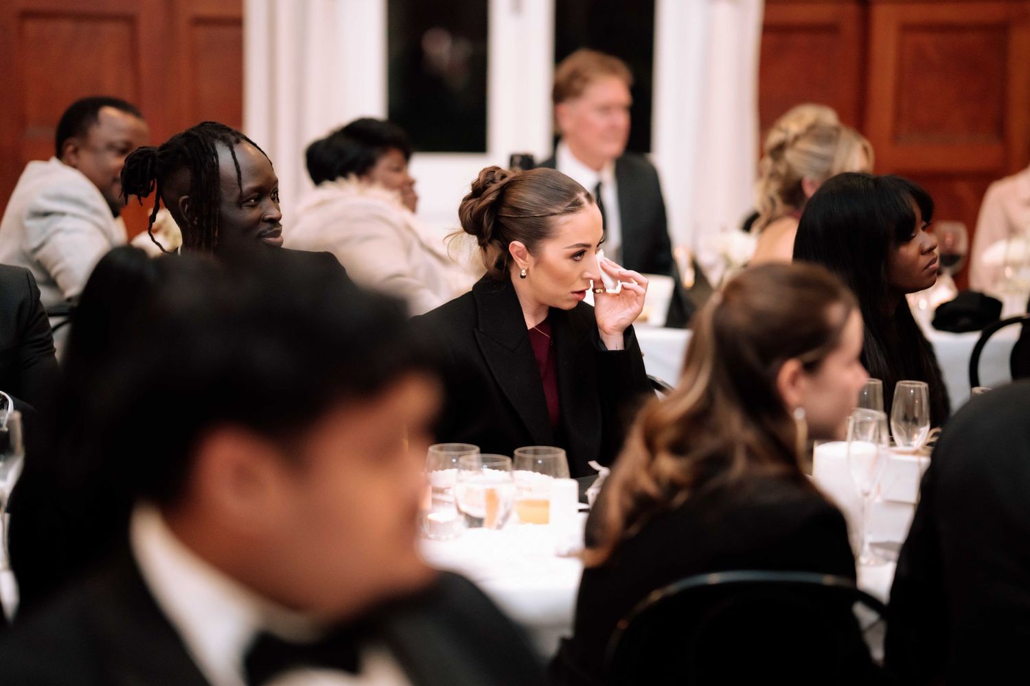 Elegantly dressed wedding reception guests seated at white-clothed dinner tables in a formal venue.