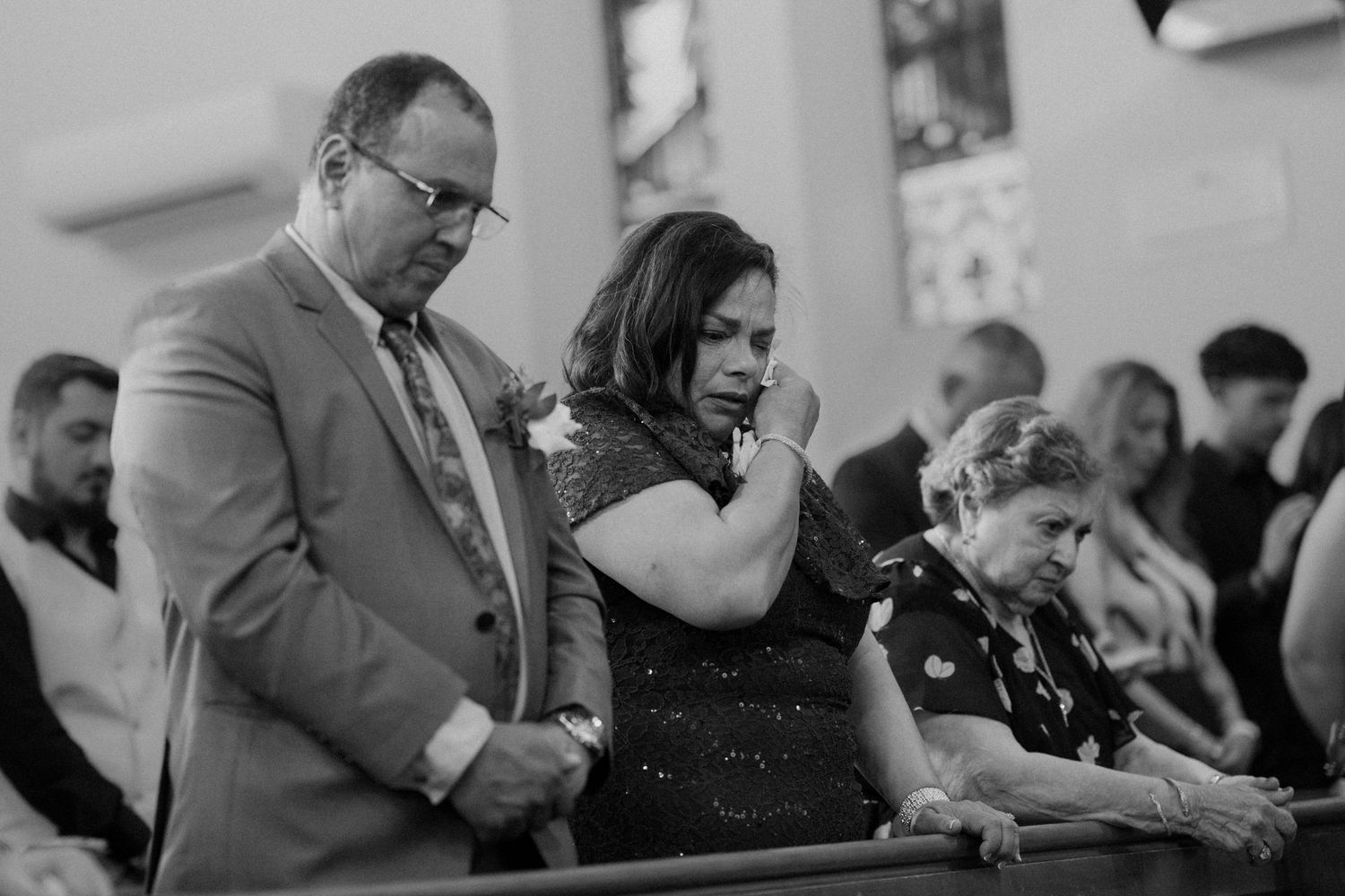 Emotional wedding guests bow in prayer during church ceremony, captured in black and white photo.