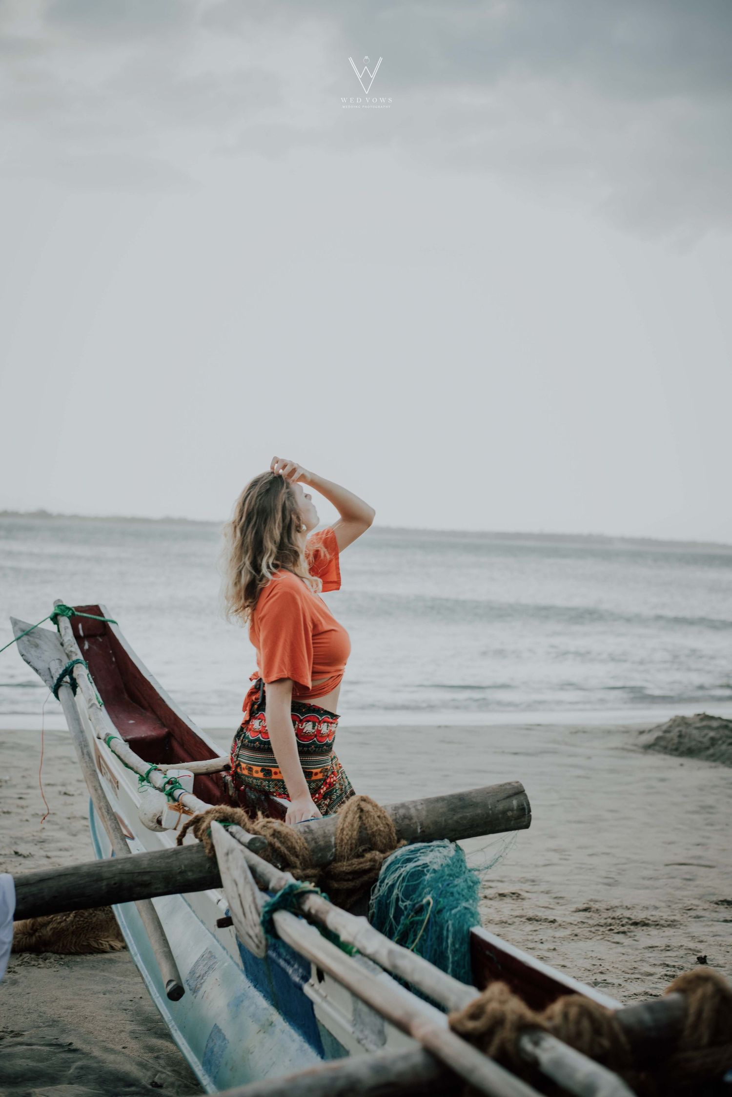 Woman in orange crop top and patterned skirt standing on a traditional wooden fishing boat on a tropical beach.