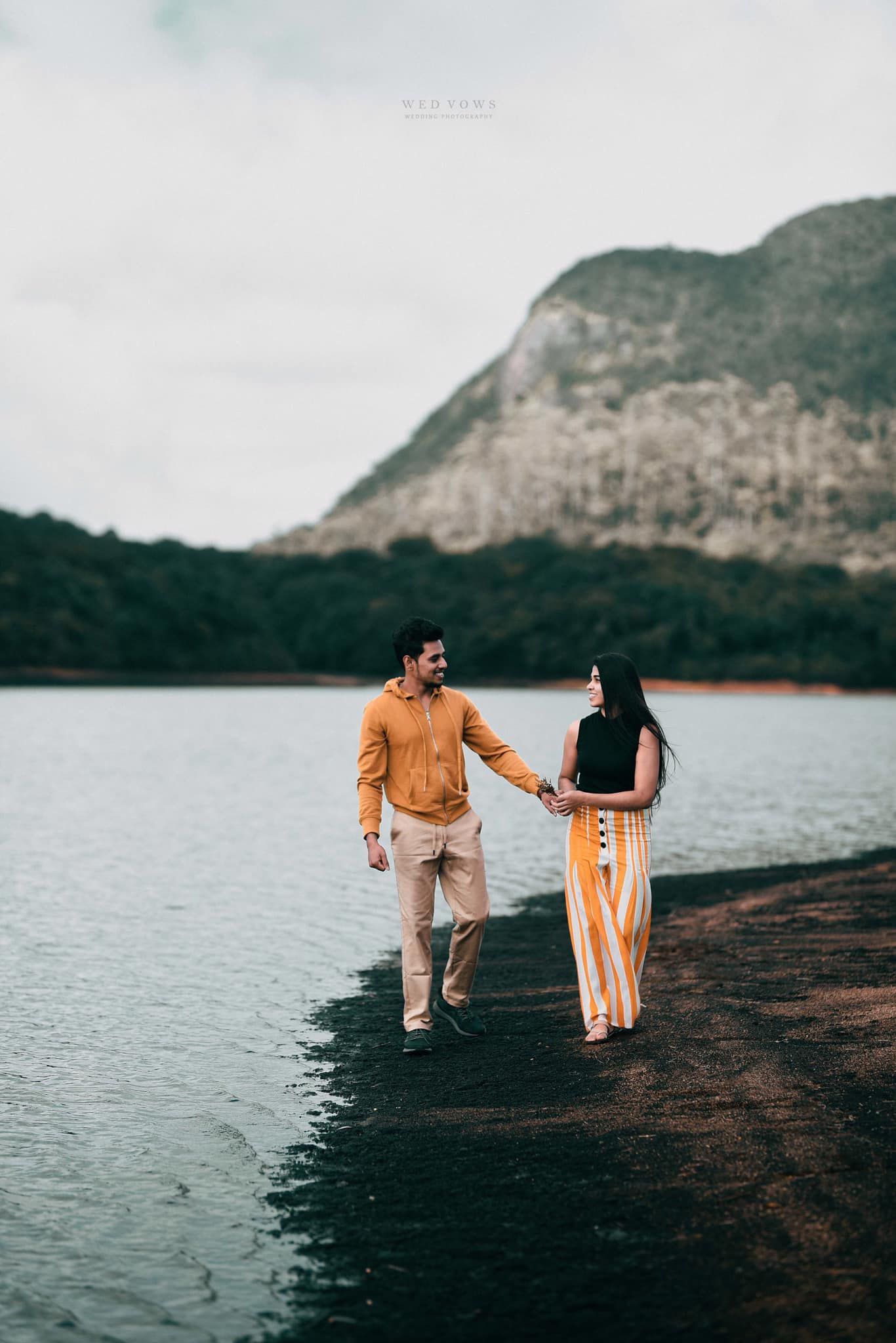 Couple holding hands and walking along rocky lakeside with mountain and cloudy sky.