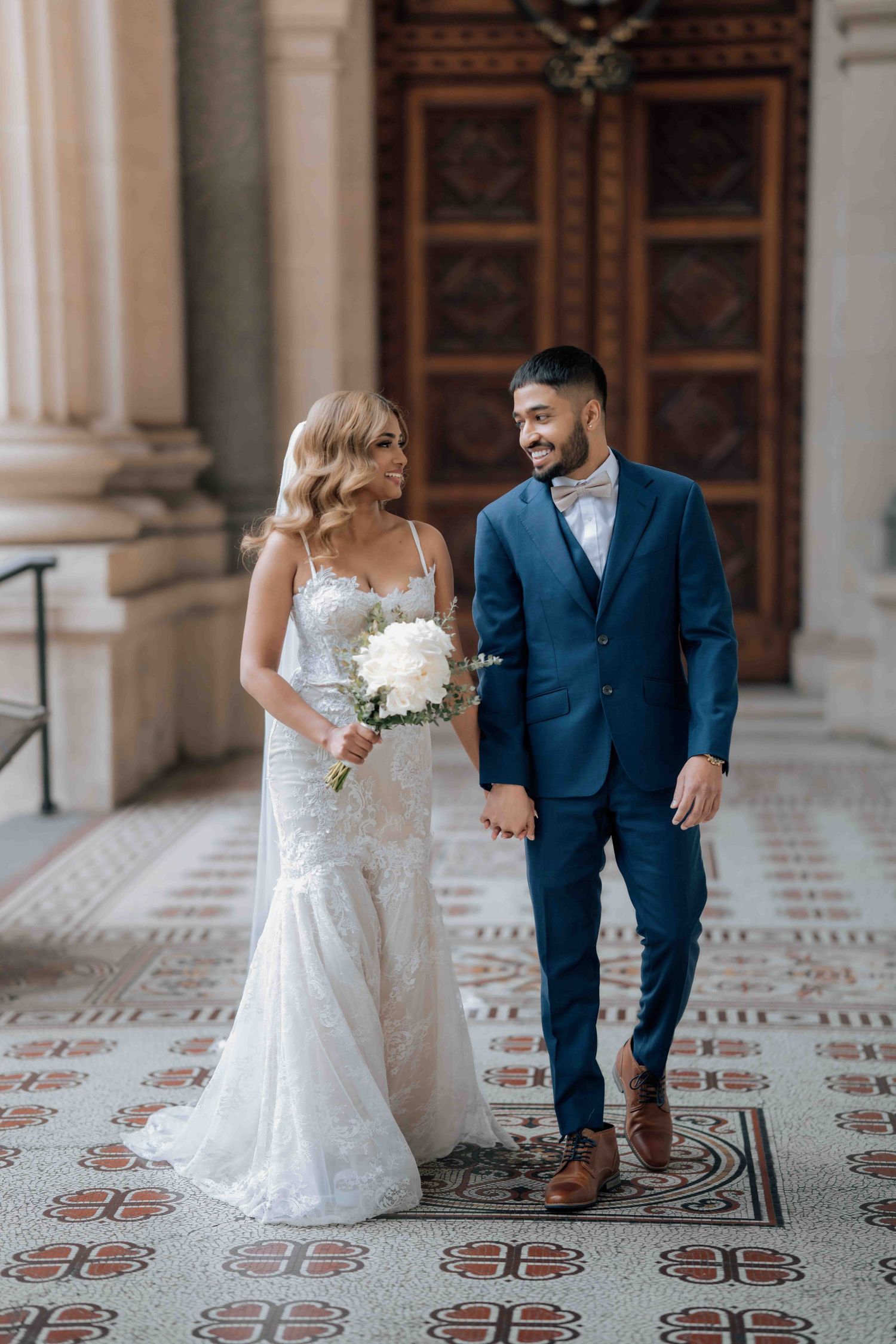 Bride and groom walking hand in hand through ornate building, bride carrying white floral bouquet.