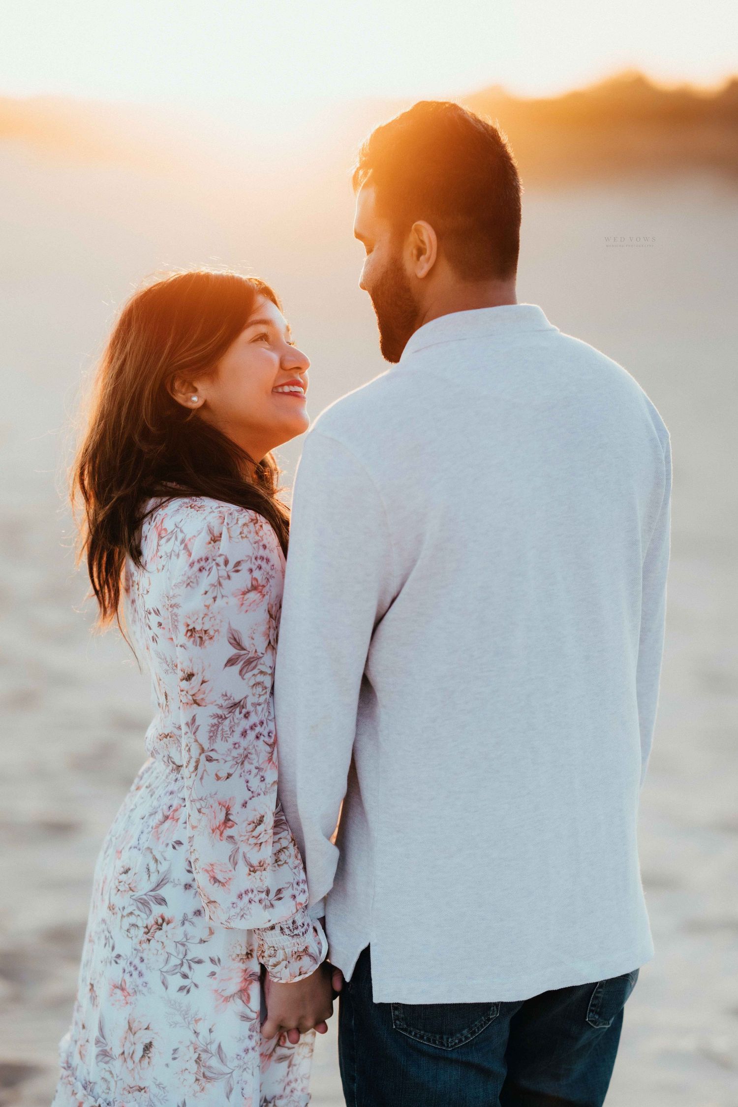 Woman smiling up at her partner while holding hands on a sunlit sandy beach at sunset.