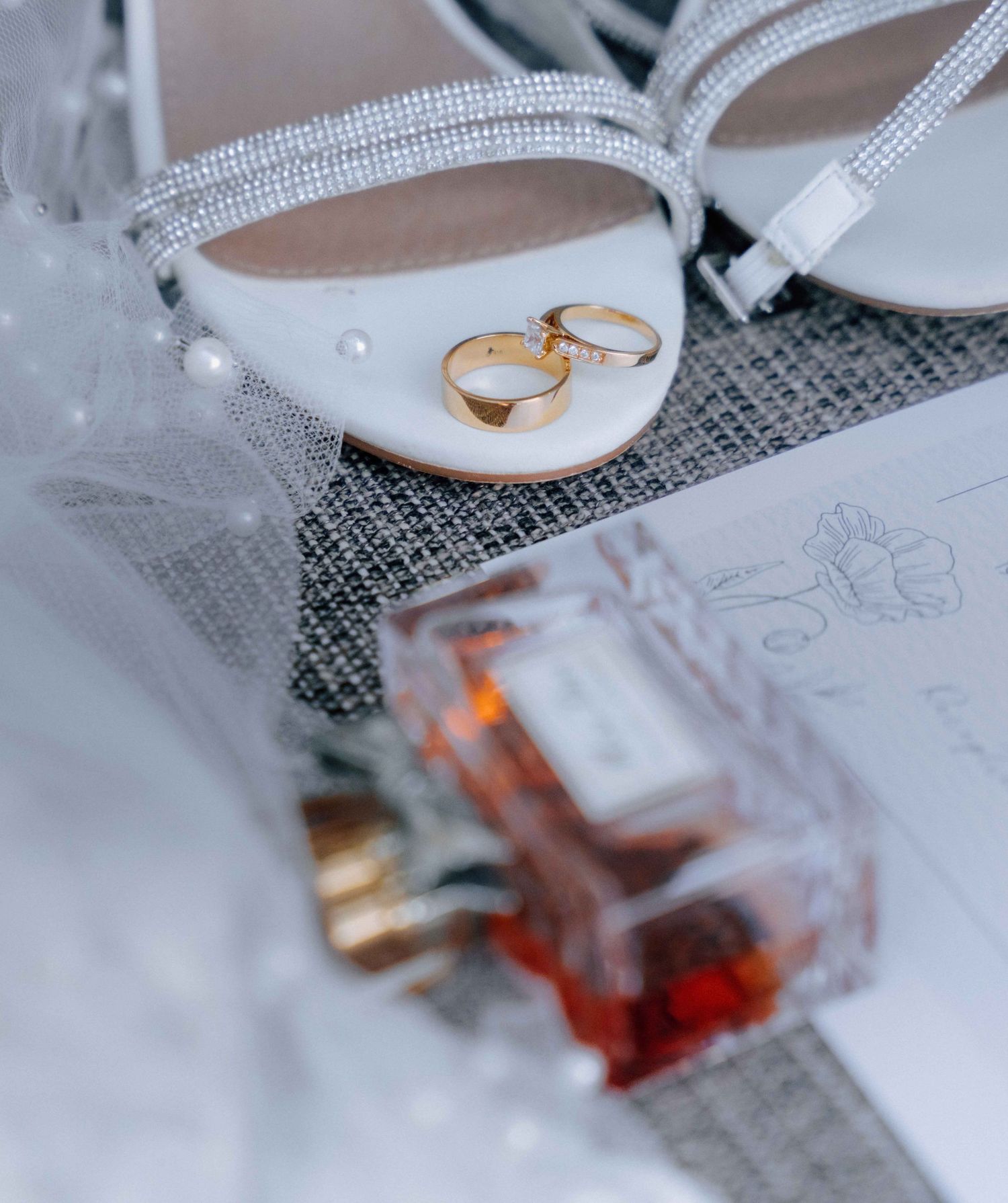 Wedding rings resting on white bridal heels beside a perfume bottle and veil on a gray surface.