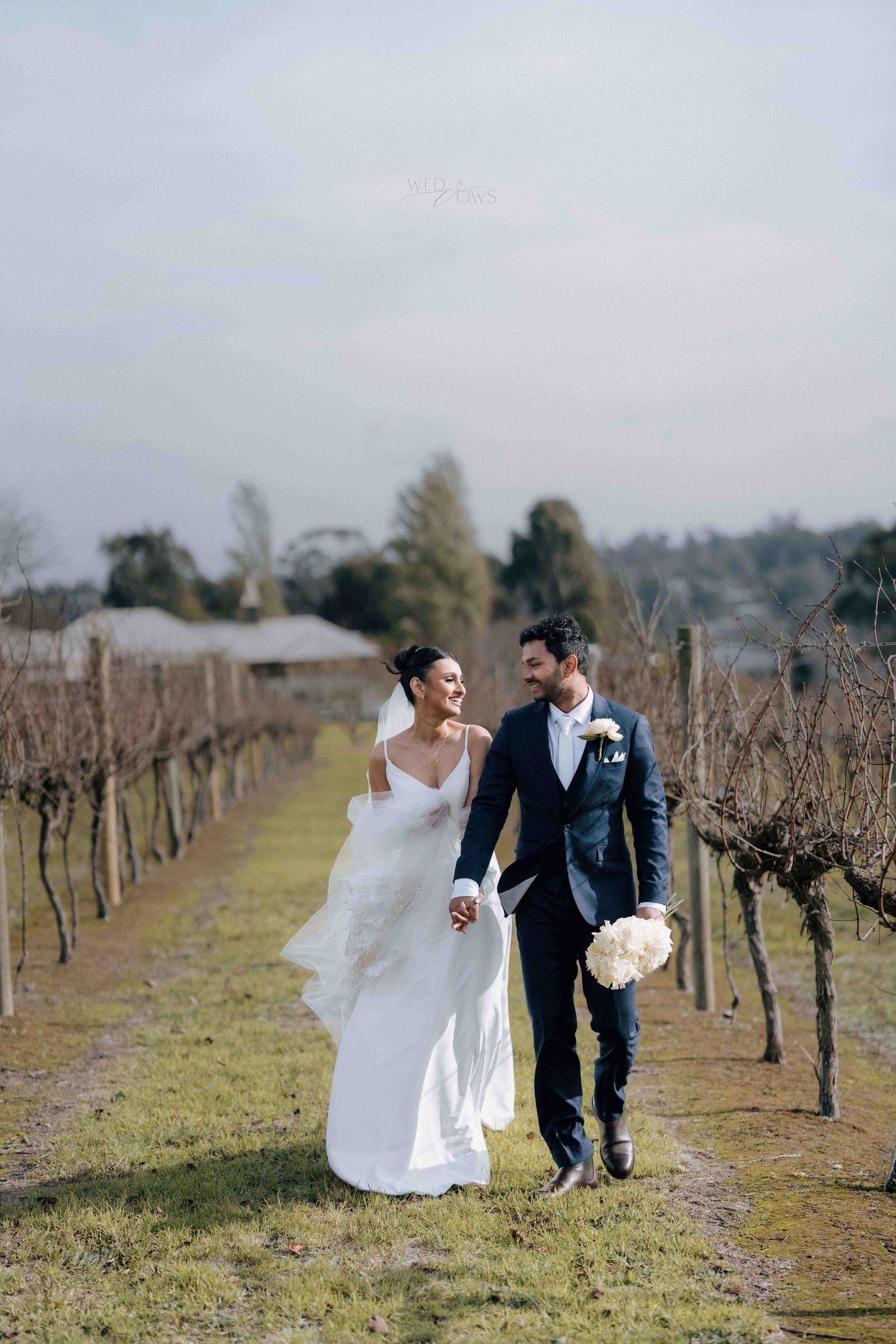 Happy bride and groom walk hand-in-hand through a winter vineyard, bride carrying white bouquet.