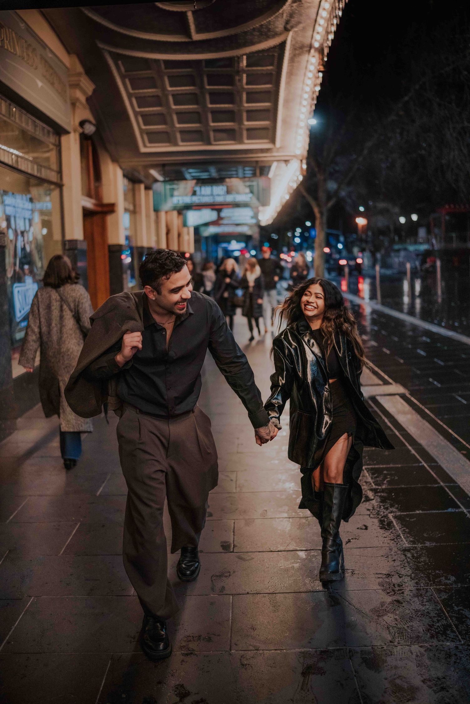 Happy couple holding hands and walking along a wet city sidewalk under bright theater lights at night.