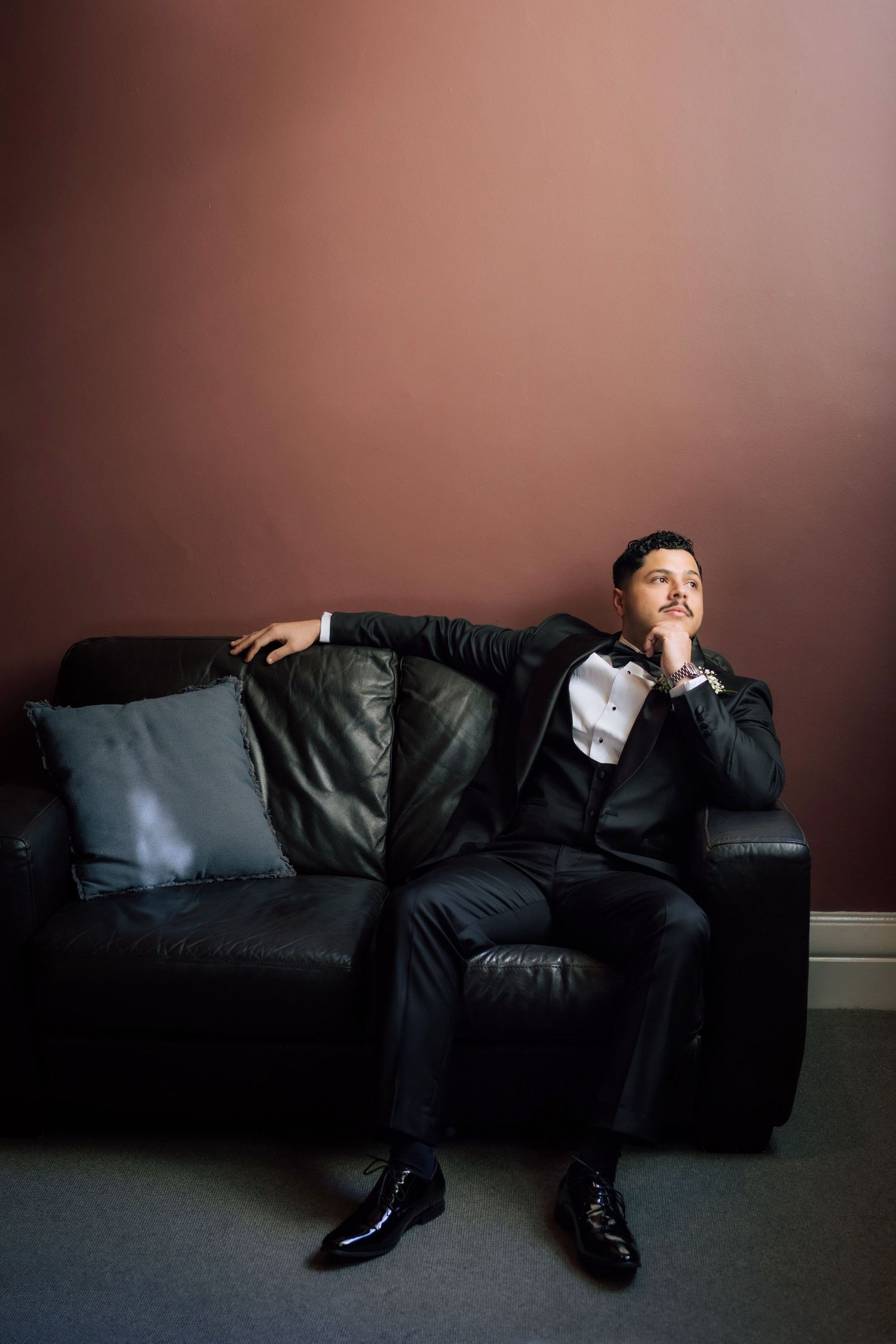 Dapper groom in black tuxedo sits relaxed on leather couch gazing upward in moody pre-wedding portrait.