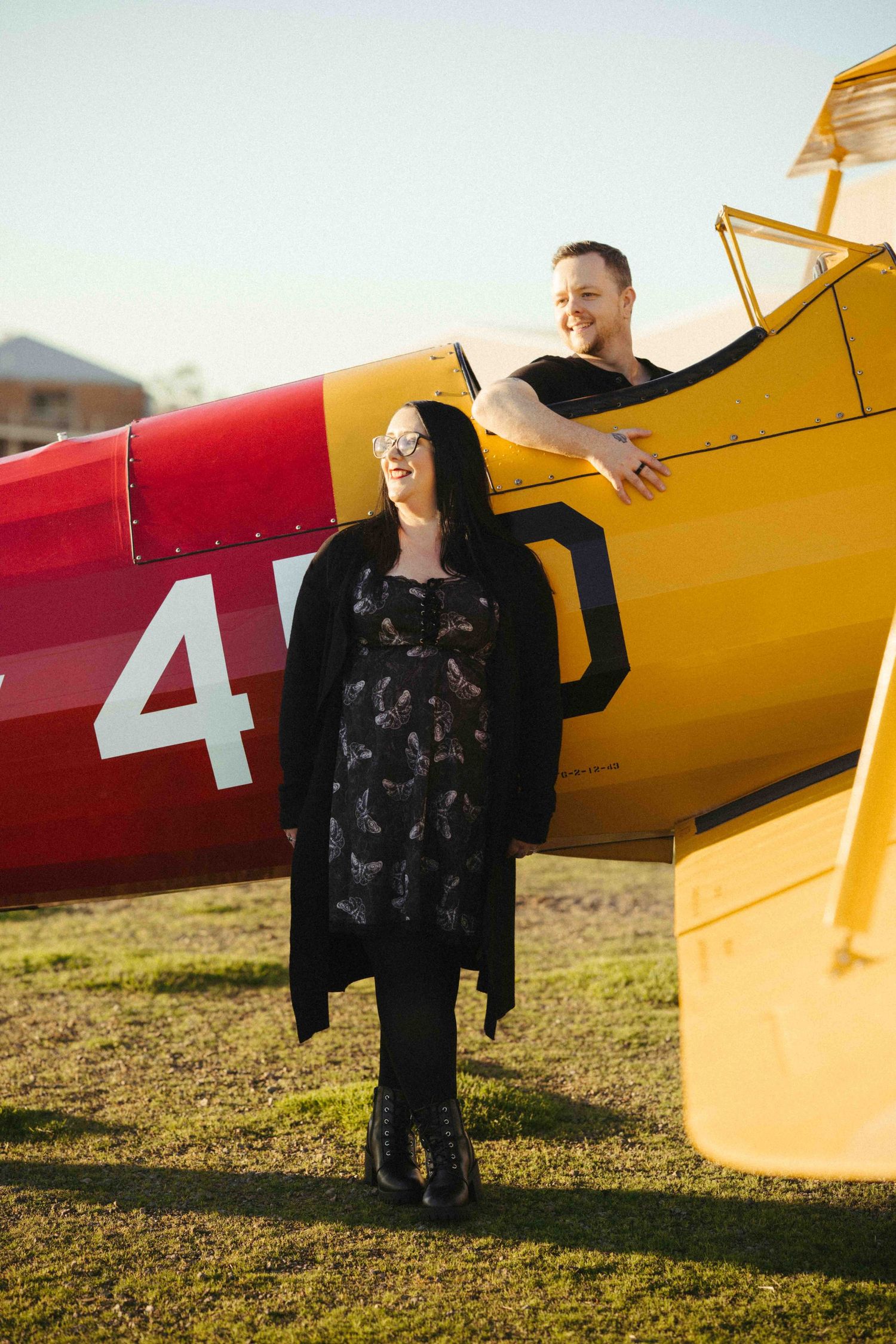 A woman in a black dress stands beside a vintage yellow and red biplane while a man smiles from the cockpit.