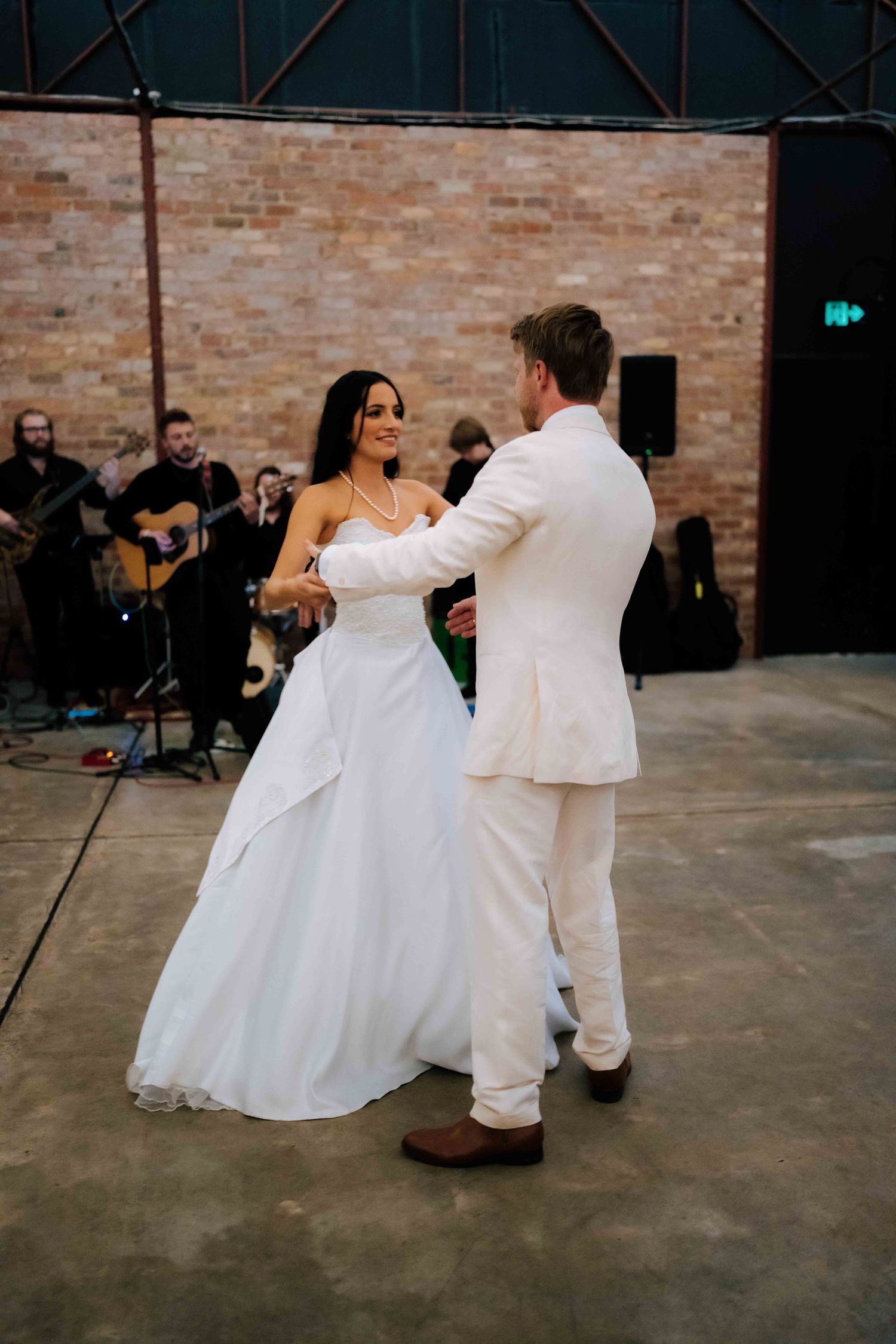 Bride and groom sharing first dance in rustic barn venue with live band performing in background.