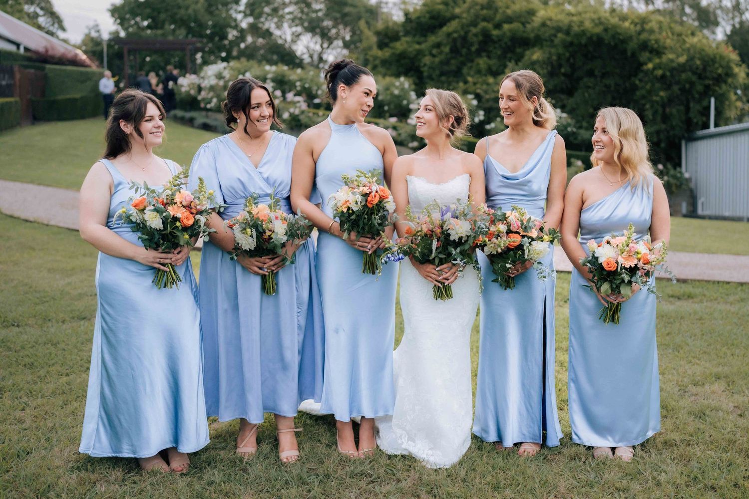 Five bridesmaids in light blue satin dresses holding colorful bouquets laughing together on green lawn.