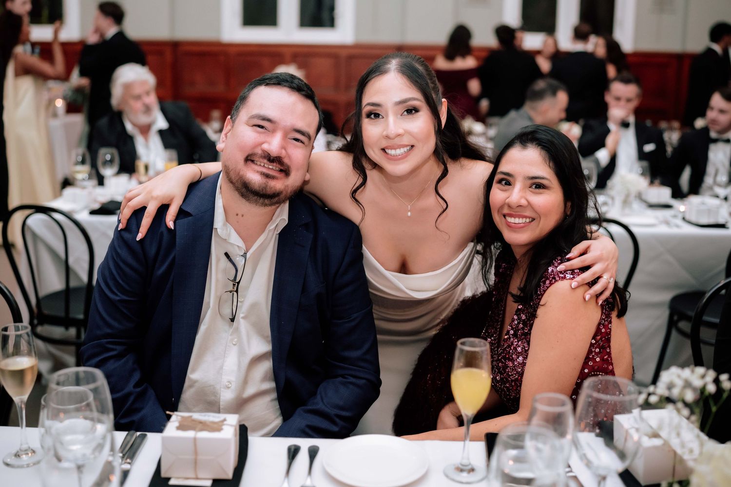 Three smiling wedding guests pose together at elegantly set reception dinner table with champagne glasses.