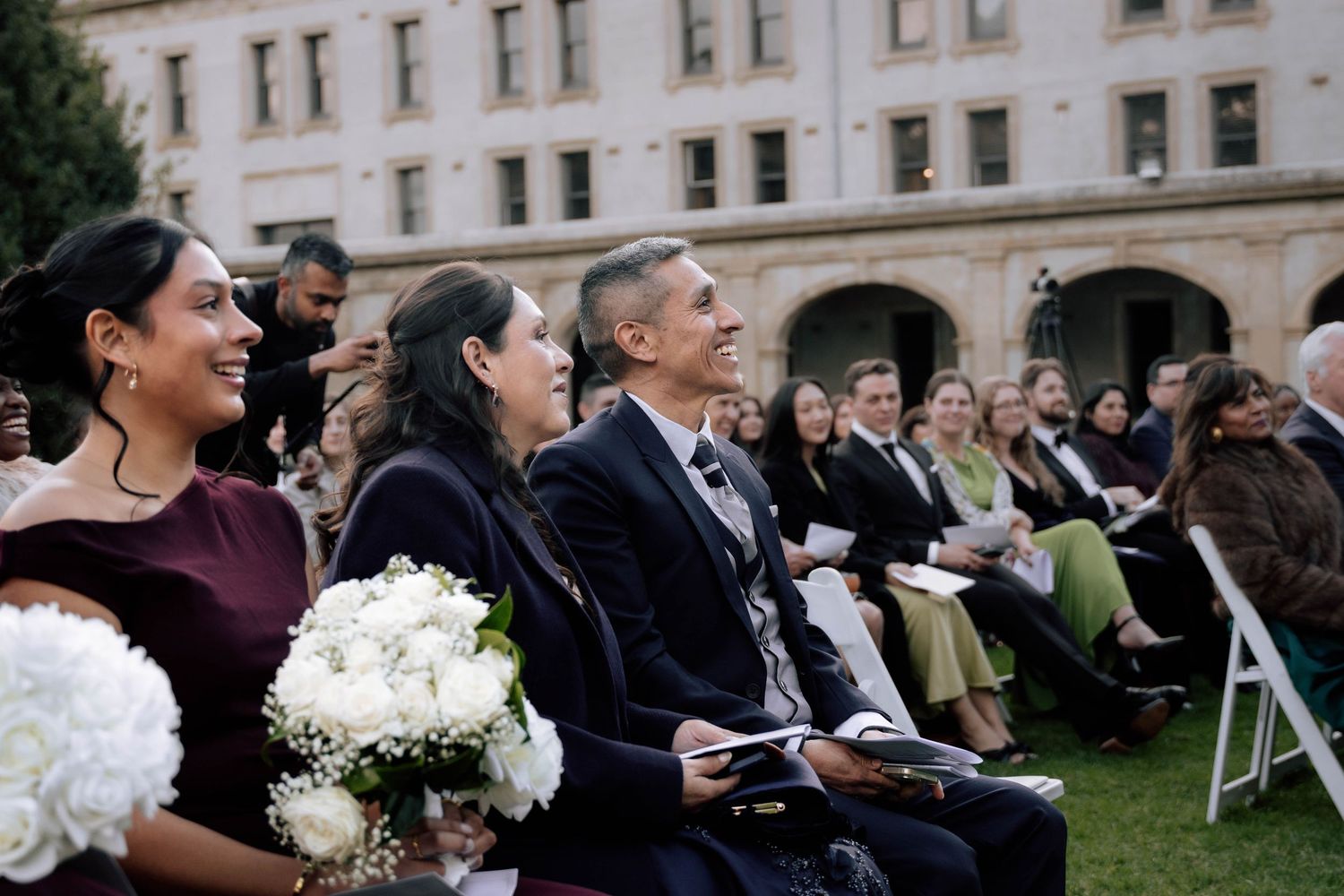 Wedding guests smiling and laughing during outdoor ceremony, bridesmaids holding white floral bouquets.