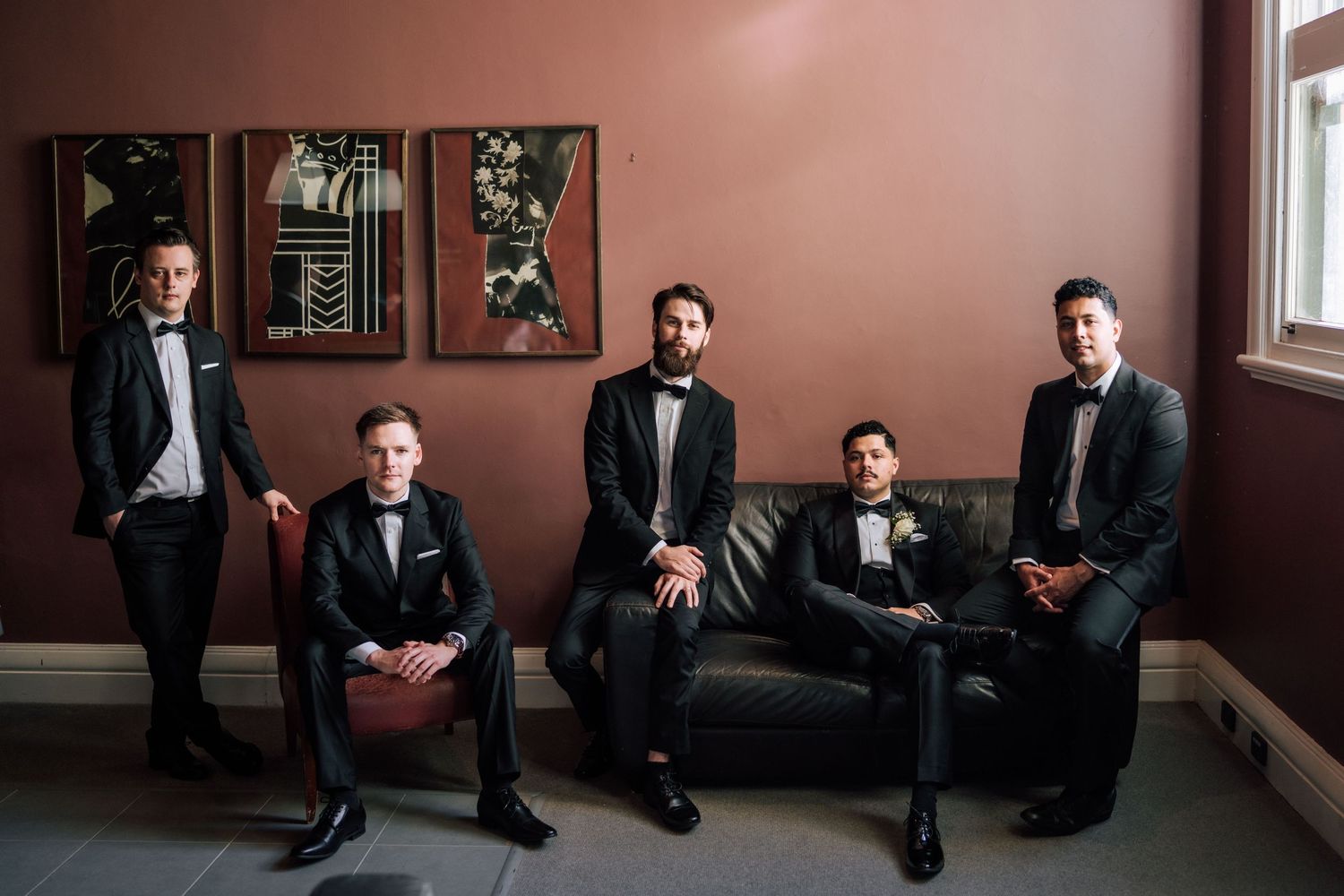 Five groomsmen in black tuxedos pose elegantly in a moody room with pink walls and vintage artwork.
