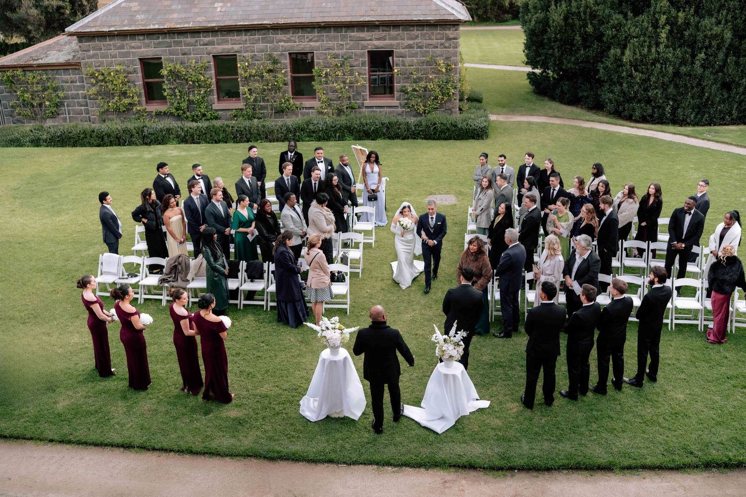 Aerial view of wedding party and guests gathered in circle on green lawn after outdoor ceremony.