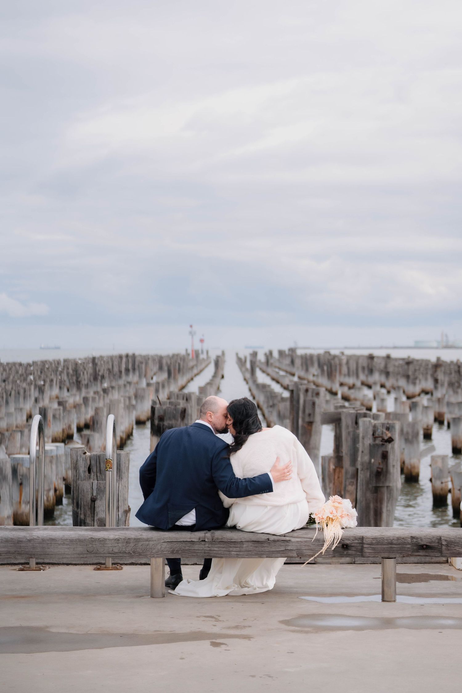 Bride and groom sharing a kiss while seated on wooden pier overlooking calm ocean waters on their wedding day.