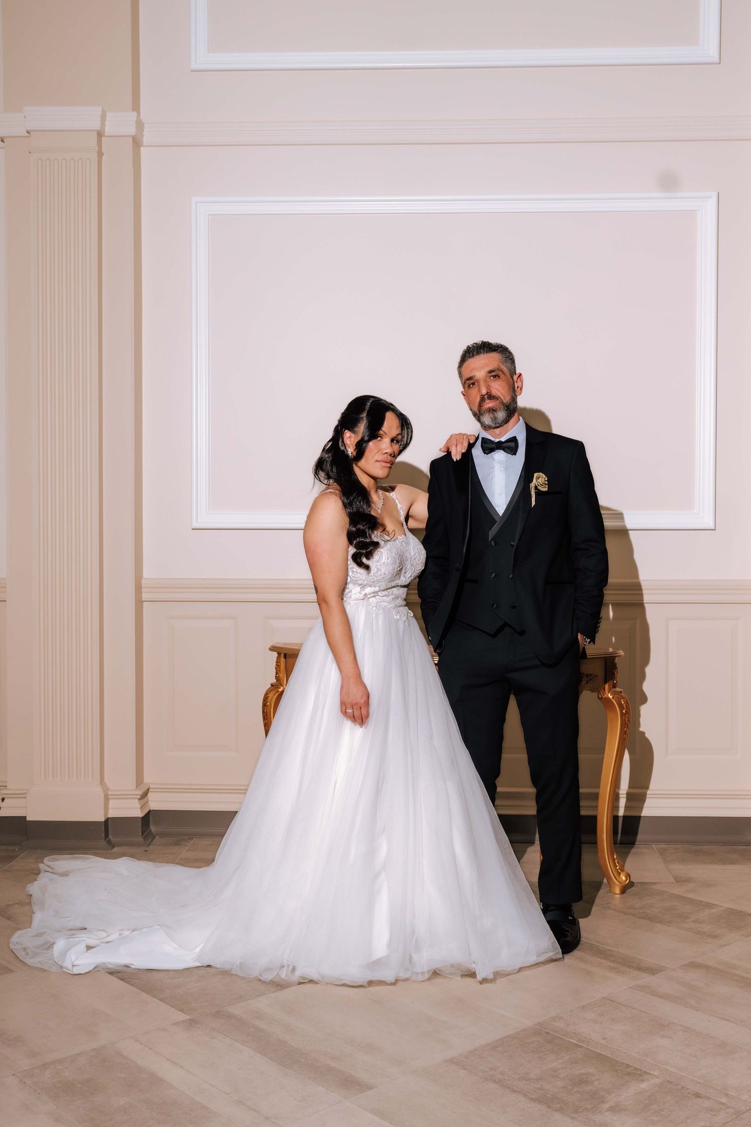 Bride and groom posing together in an elegant ballroom, bride in white tulle gown and groom in black suit.