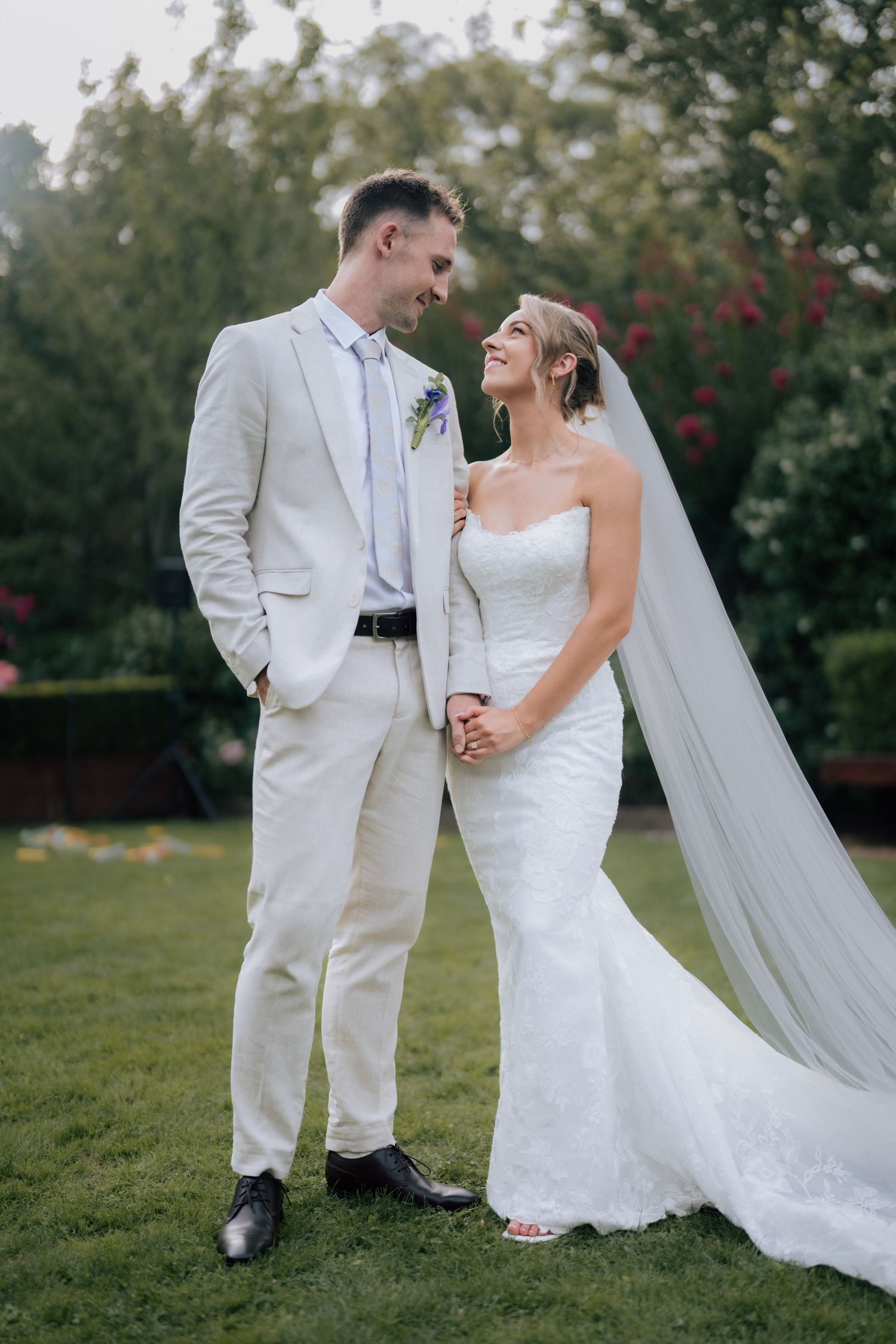 Bride in strapless lace gown and groom in cream suit holding hands and smiling in garden setting.