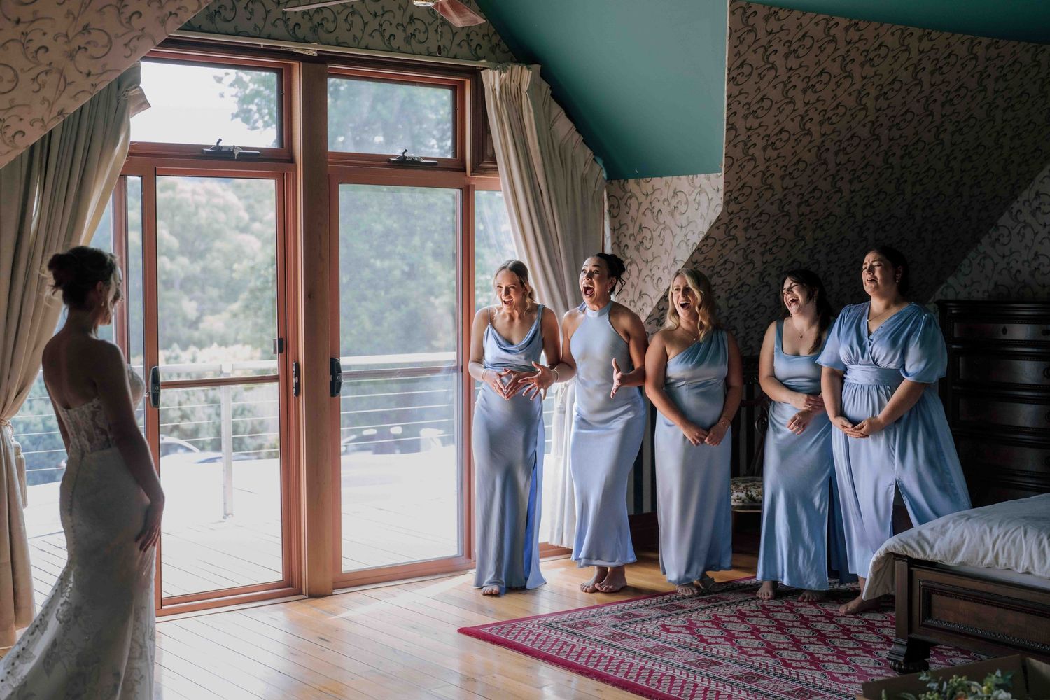 Bridesmaids in blue dresses react emotionally to bride's first look in bright room.