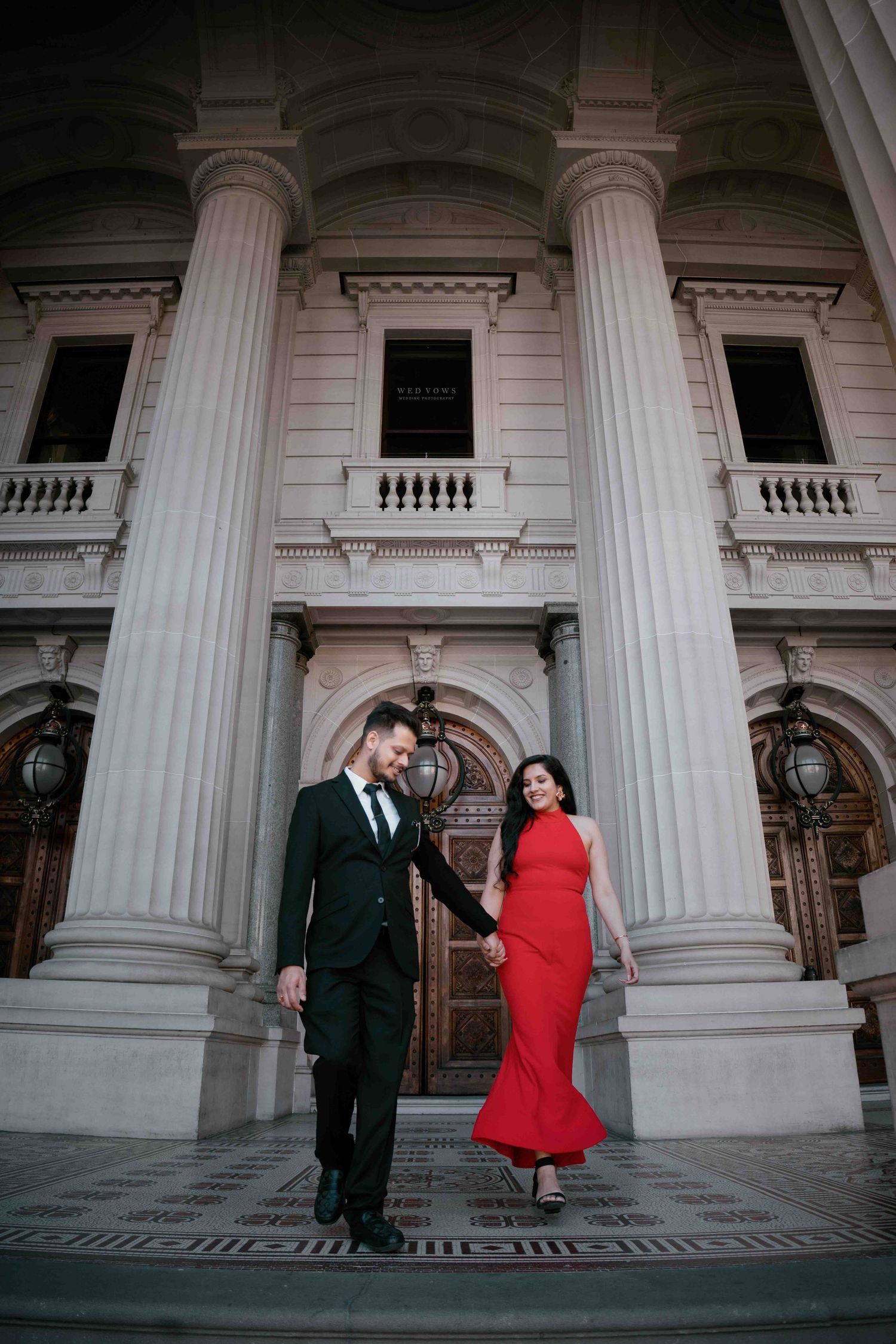 Elegant couple in formal wear walking hand in hand before grand neoclassical building.