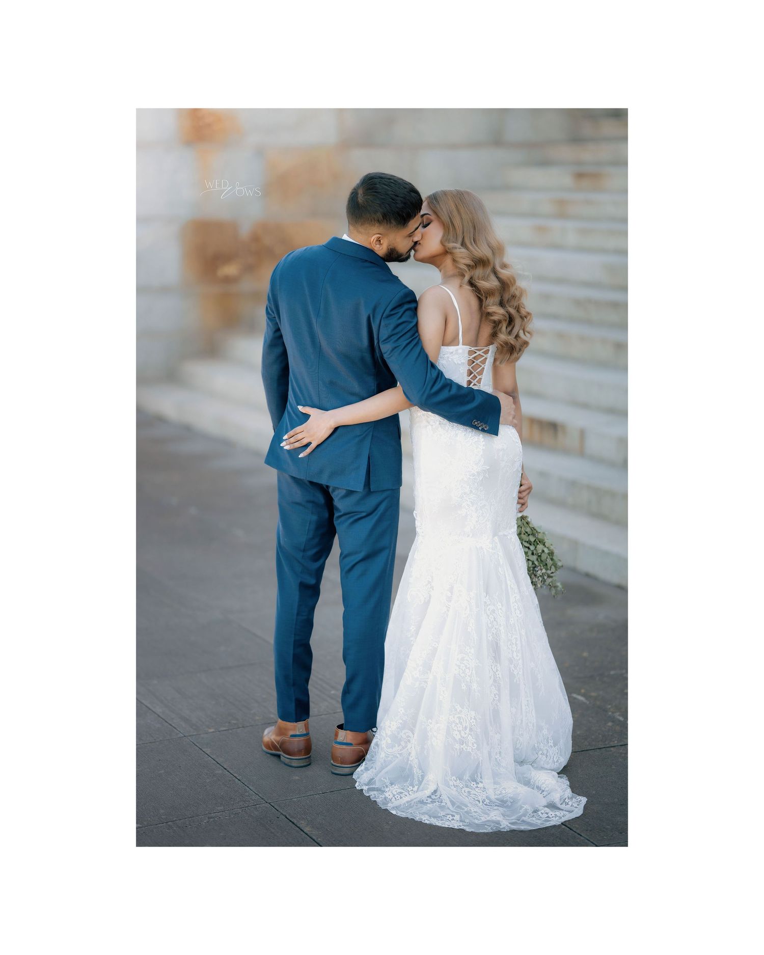 Couple embracing and kissing, bride in backless lace gown with train, groom in navy suit.