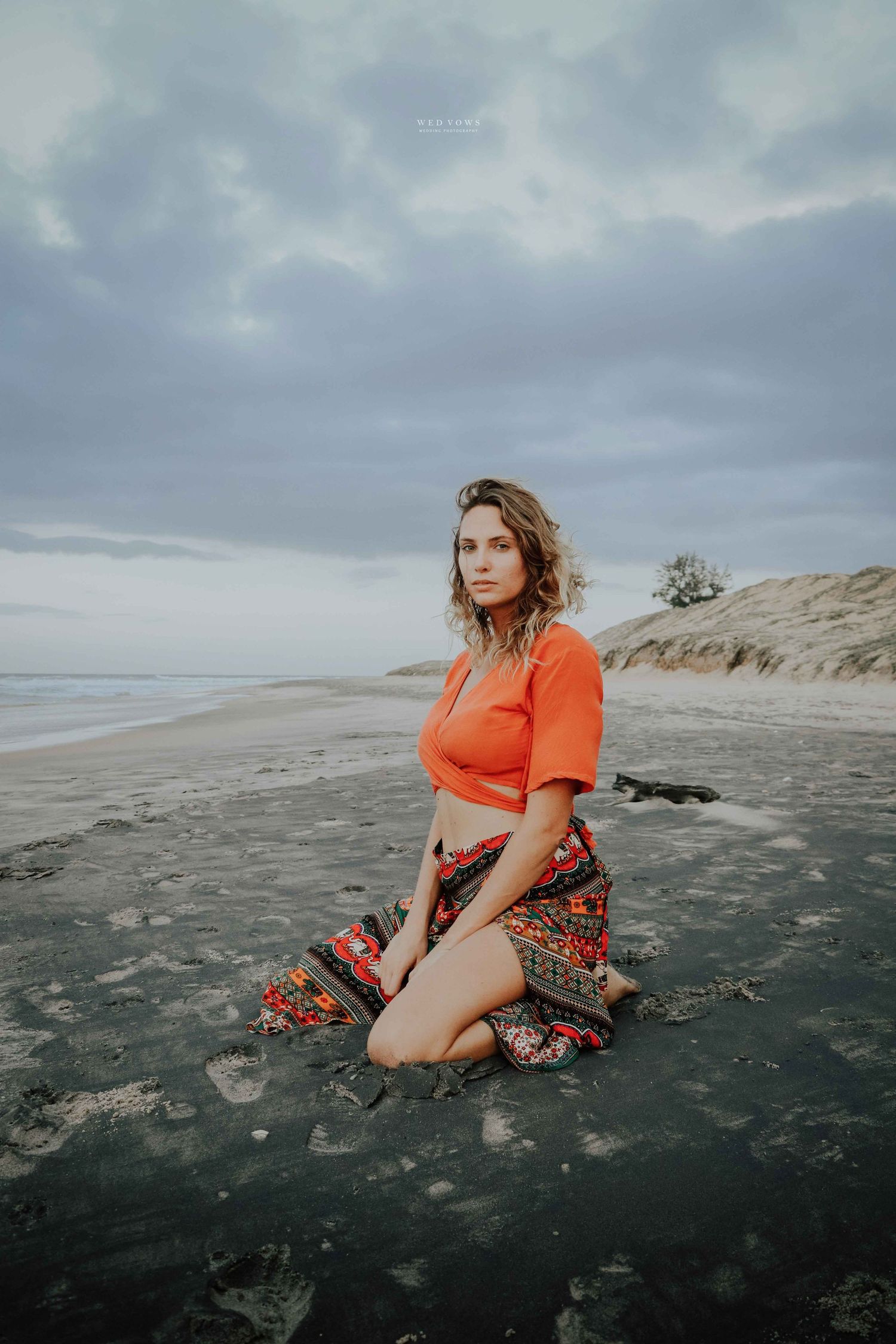 Woman in orange crop top and bohemian skirt kneeling on dark wet sand with cloudy sky.