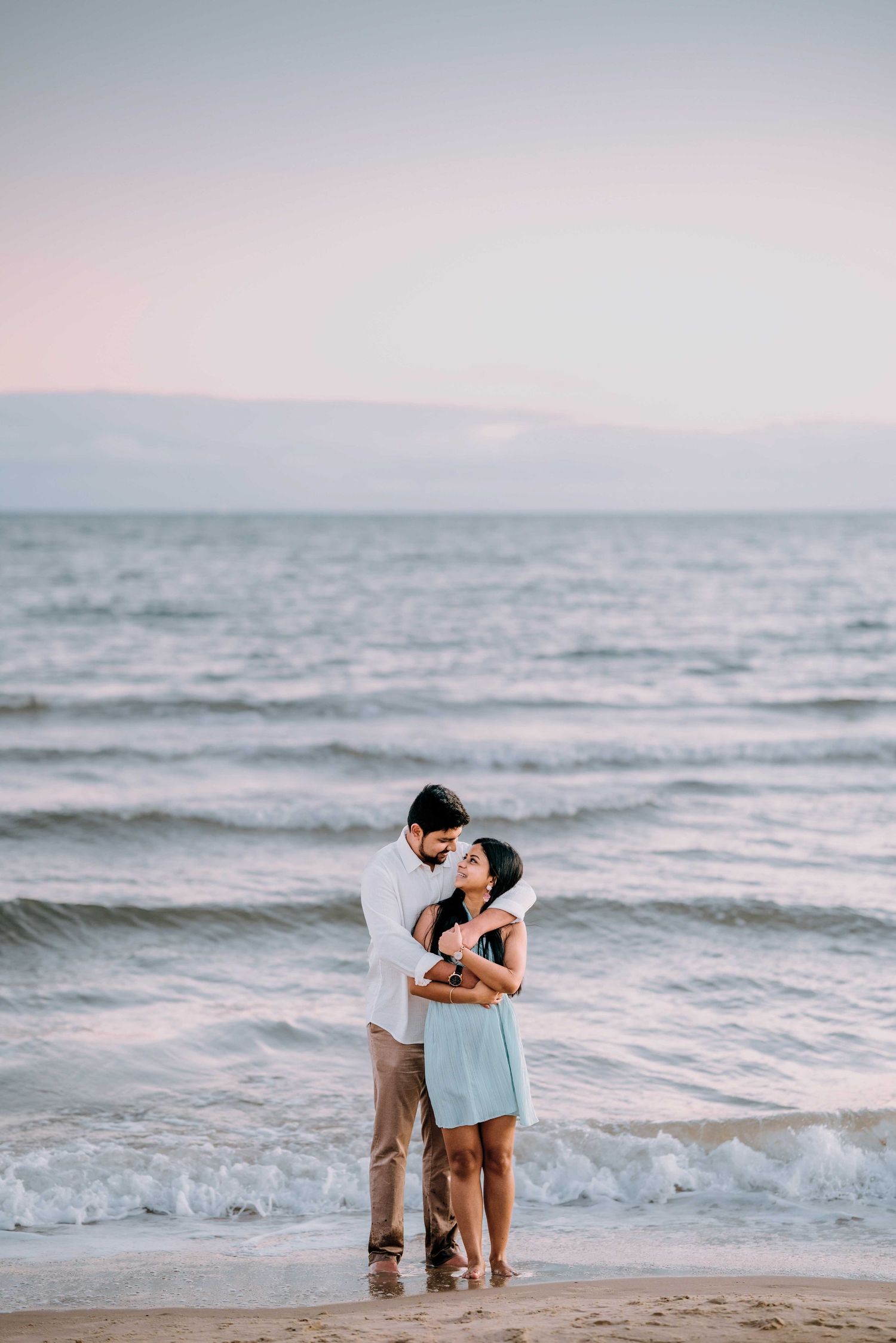 Loving couple embracing on sandy beach at dusk, woman in light blue dress, waves in background.