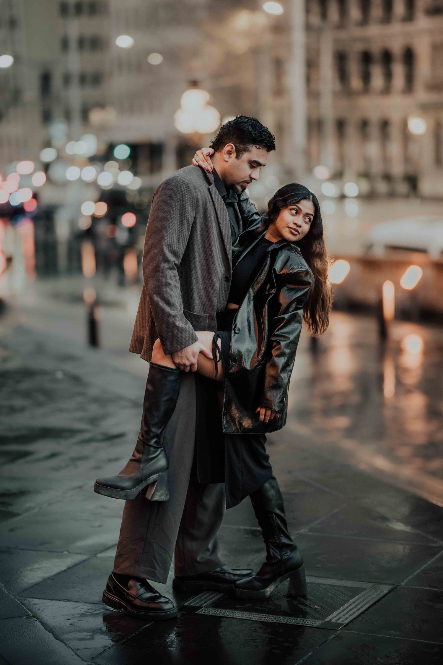 Stylish couple posing together on a rain-soaked city street at night with bokeh lights.