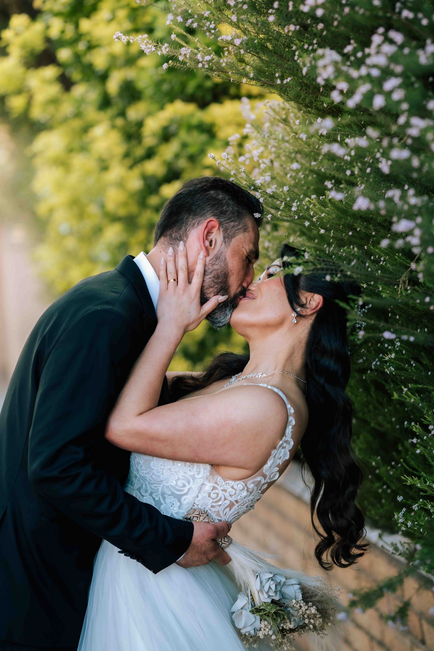 A bride and groom share a romantic kiss surrounded by blooming white and green foliage on their wedding day.