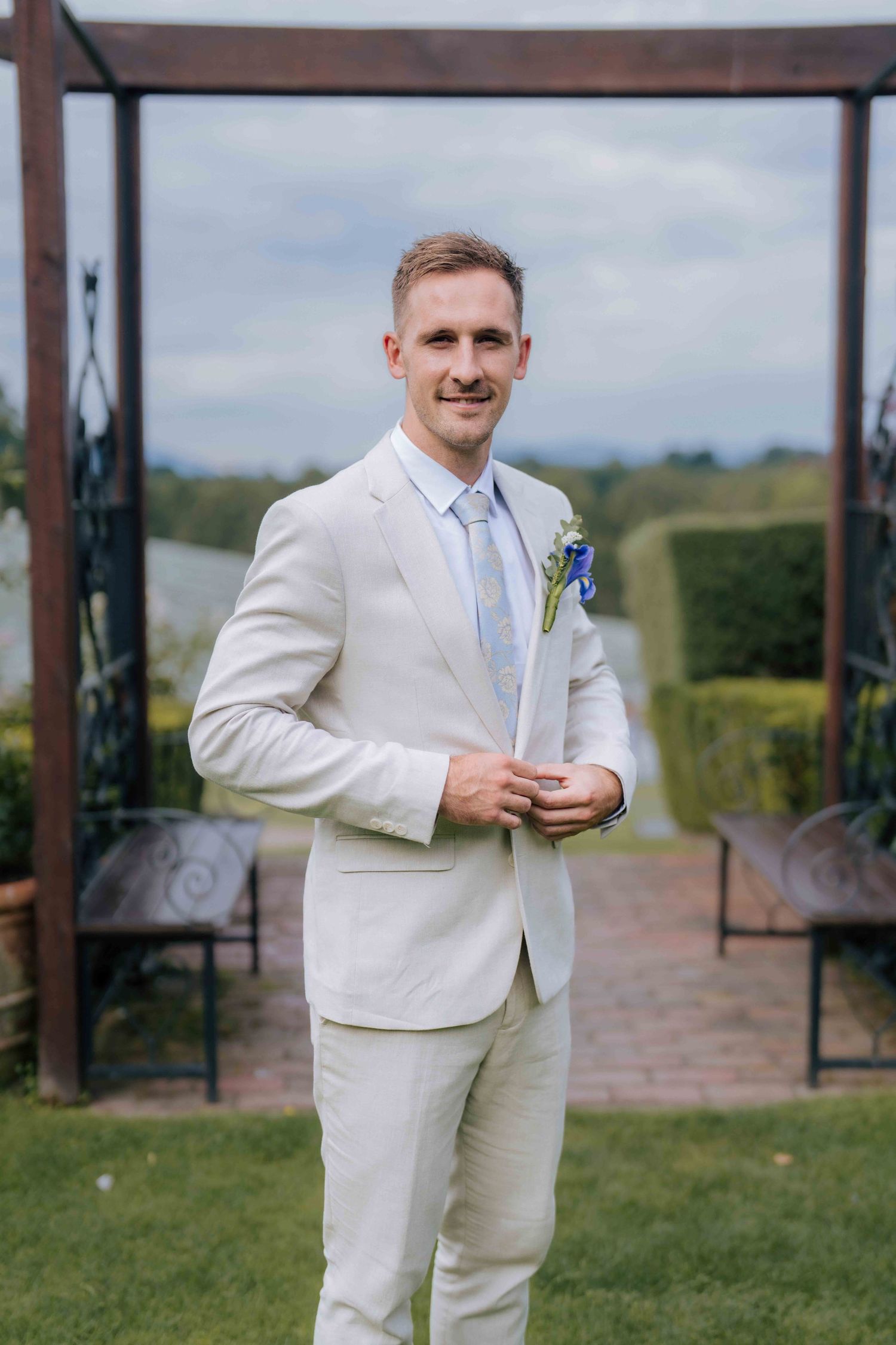 Groom in cream suit with blue boutonniere stands confidently before wooden wedding arch.