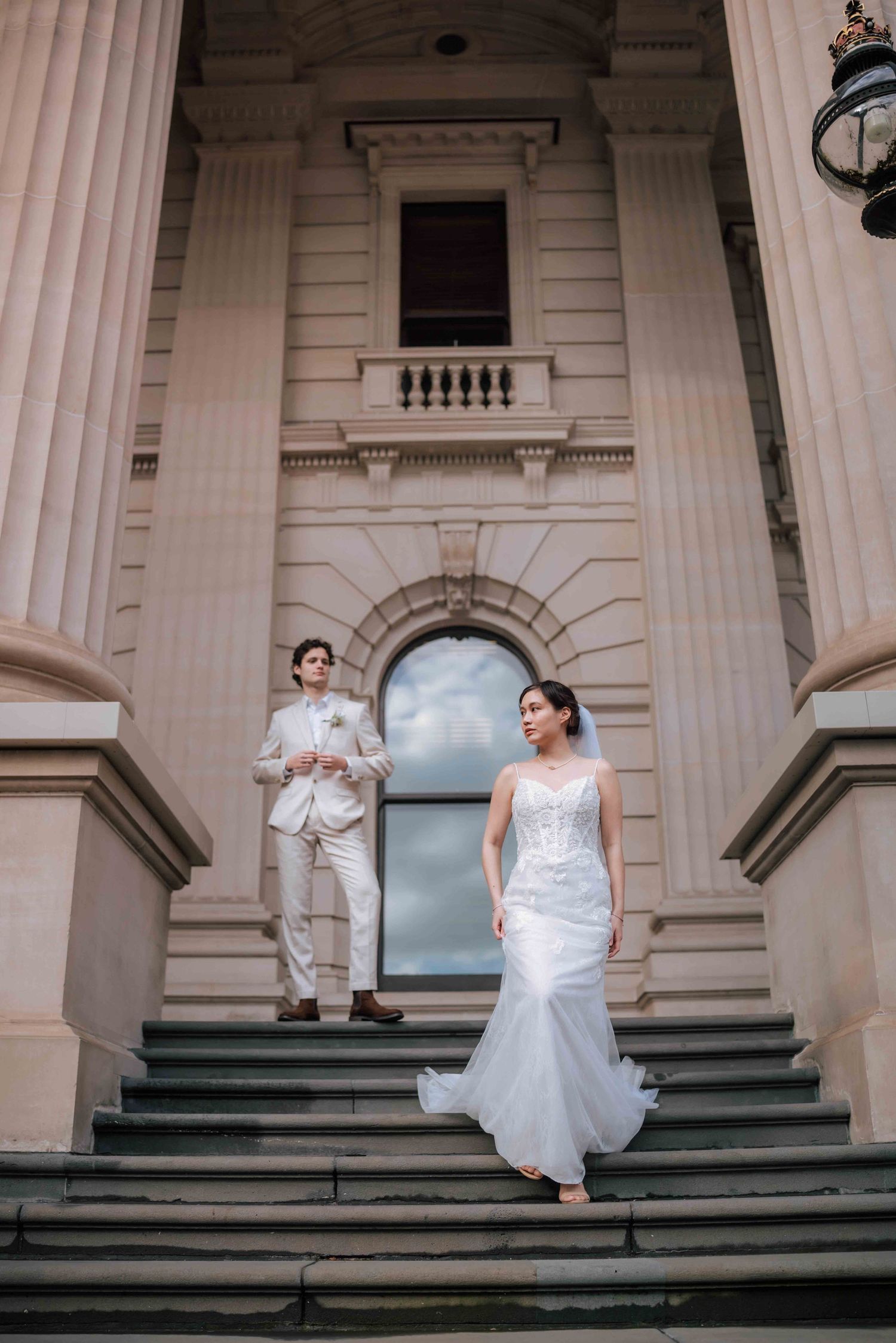 Bride in flowing lace gown and groom in beige suit descend grand stone steps of a classical heritage building.