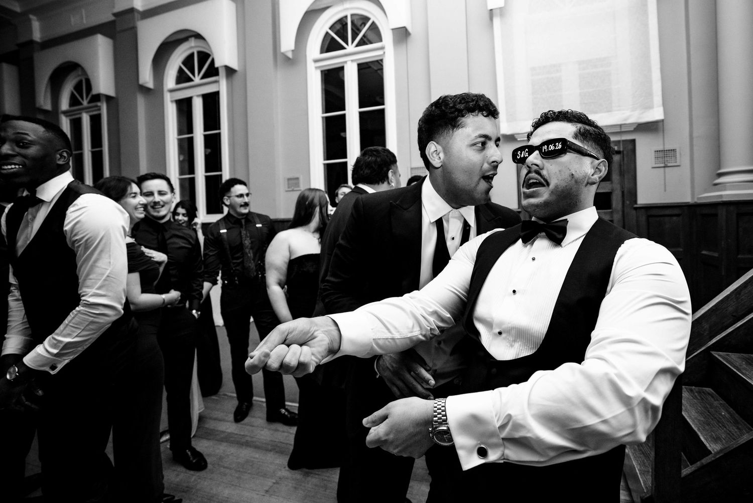 Two men in formal tuxedos dancing and celebrating together at a wedding reception.