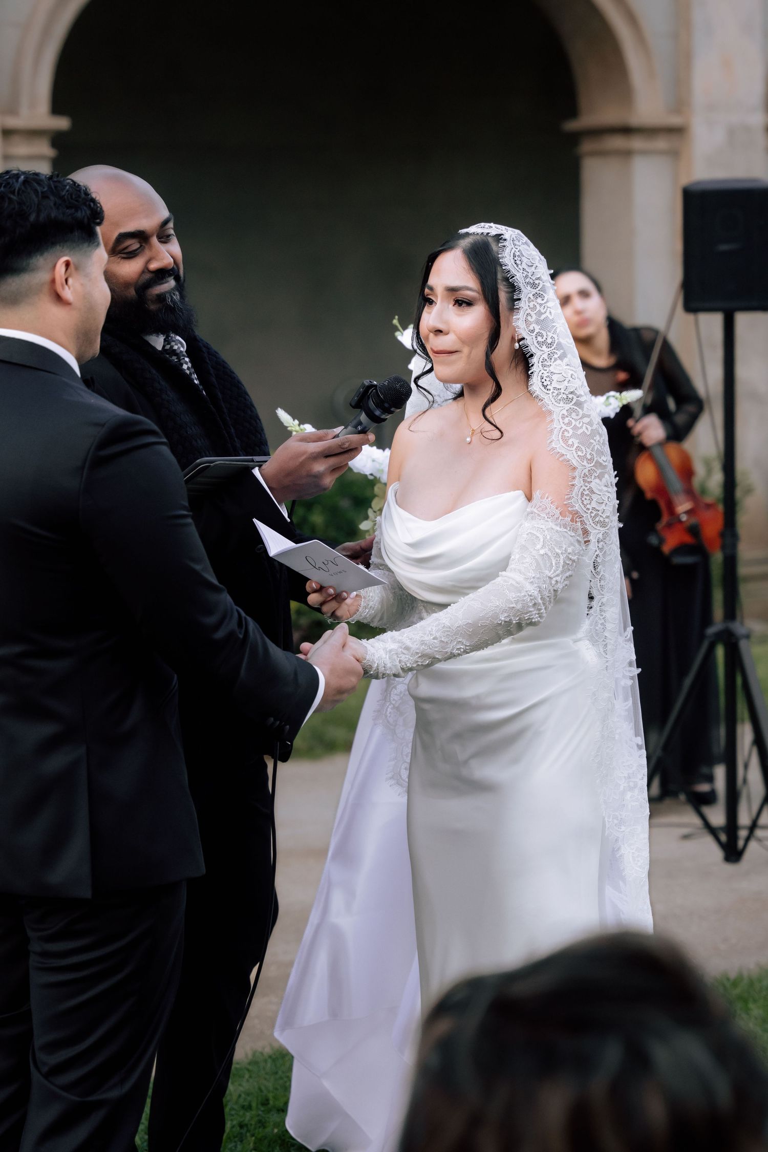 Bride in elegant white gown and lace veil exchanging vows during outdoor wedding ceremony.