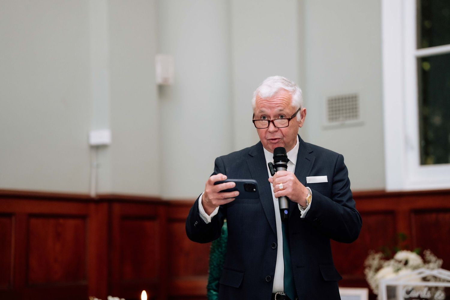 An elderly man in a suit holds a microphone and smartphone while speaking at an elegant wedding reception.