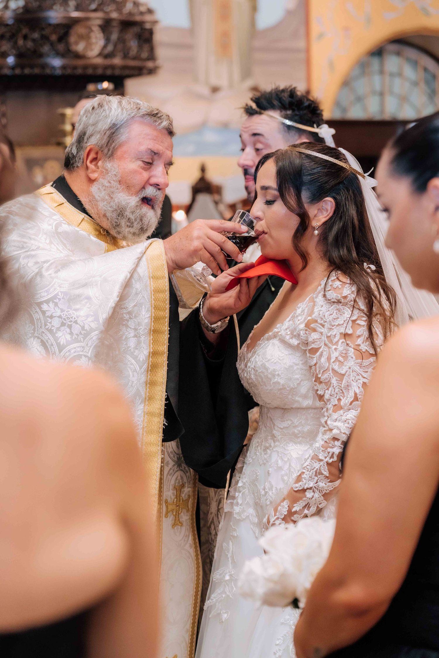 Greek Orthodox priest offers communion to bride in lace gown during wedding ceremony.