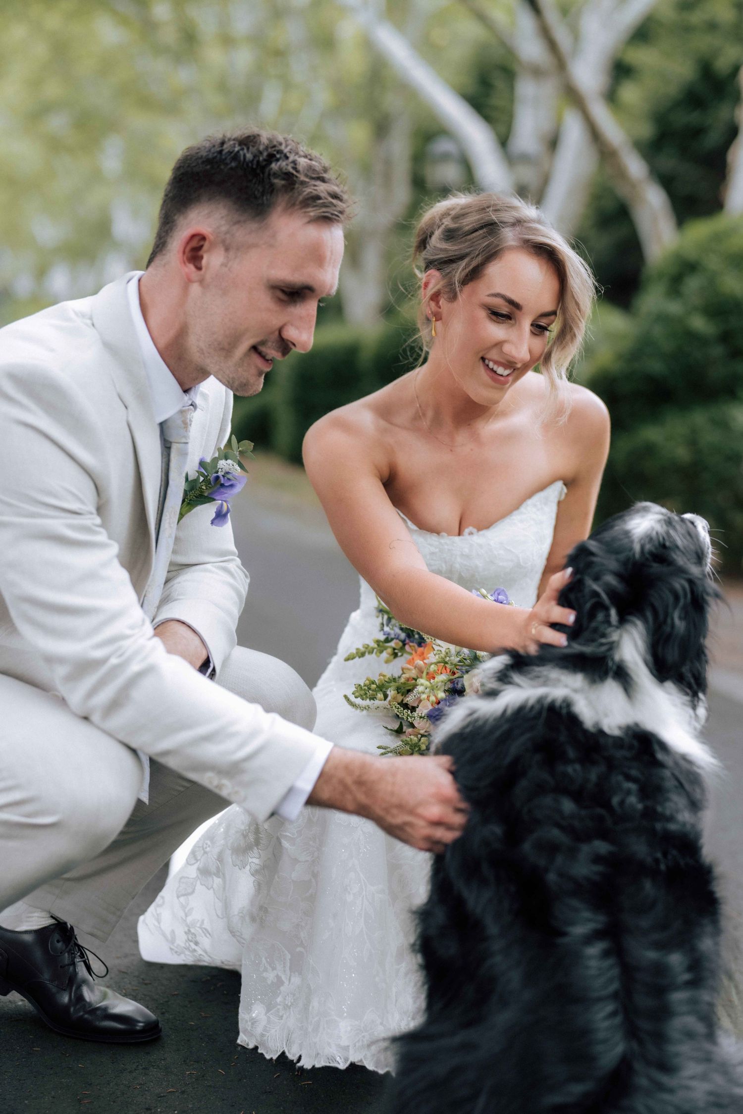 Laughing bride and groom in white attire crouching to interact with their black and white dog outdoors.