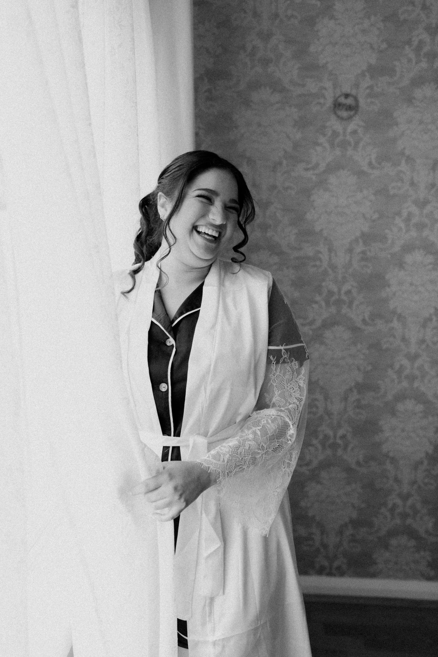 Black and white portrait of laughing bride in robe preparing by window on wedding morning.