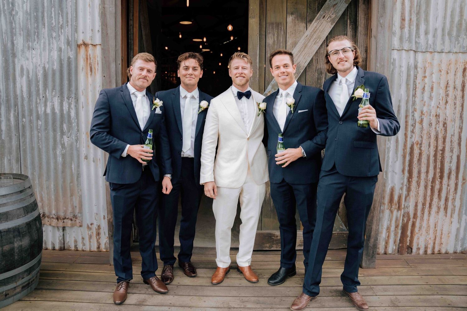 Groom in white tuxedo stands with four groomsmen in navy suits outside a rustic barn holding champagne bottles.
