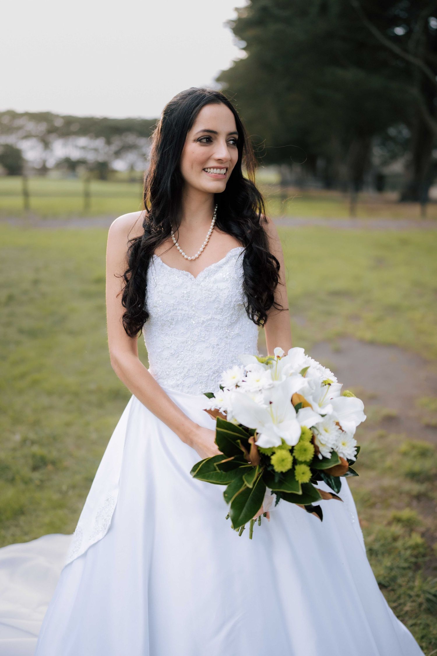 Smiling bride in white strapless gown holding bouquet of white flowers and greenery outdoors.