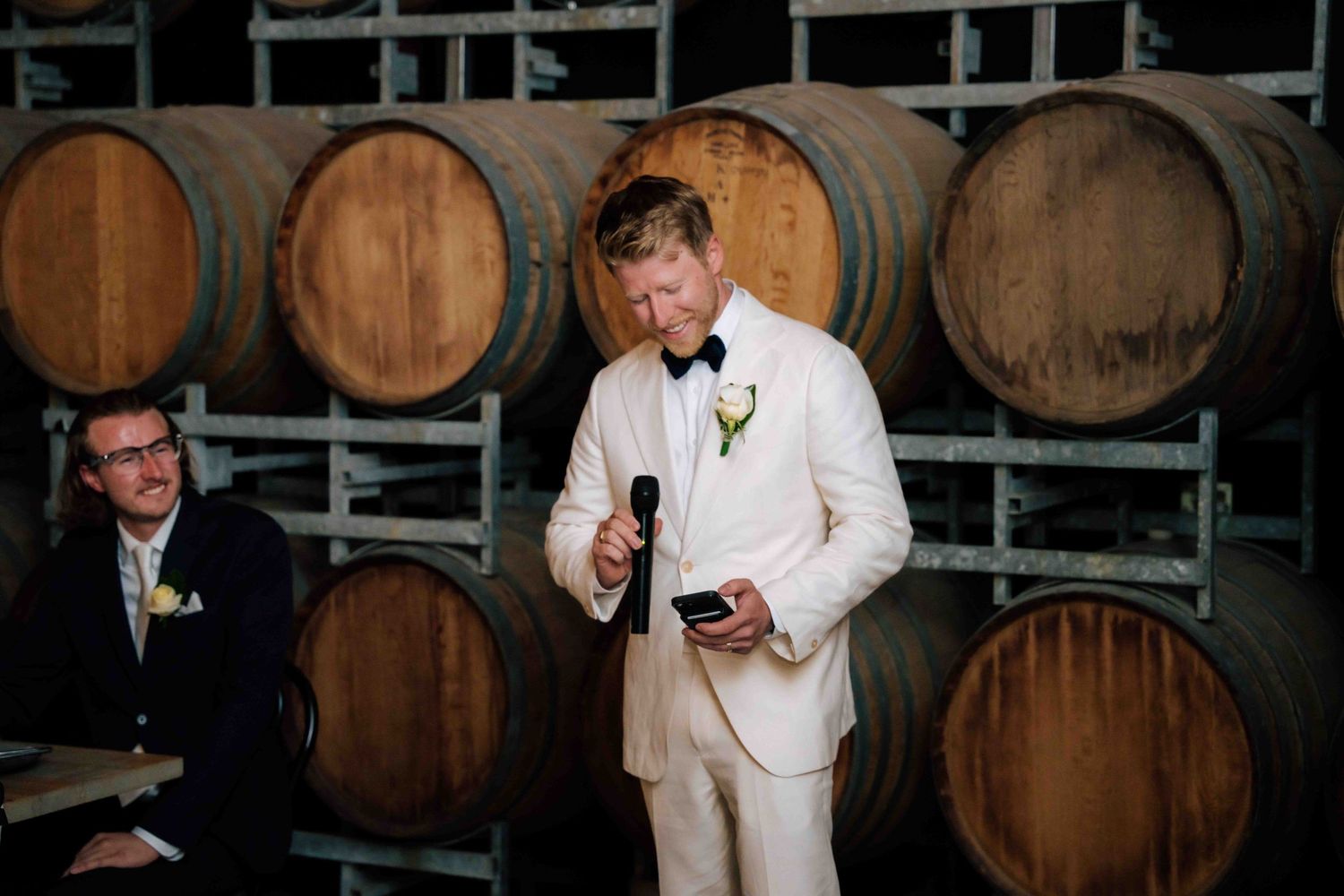 Groom in white tuxedo delivers speech near wine barrels at a winery wedding reception.
