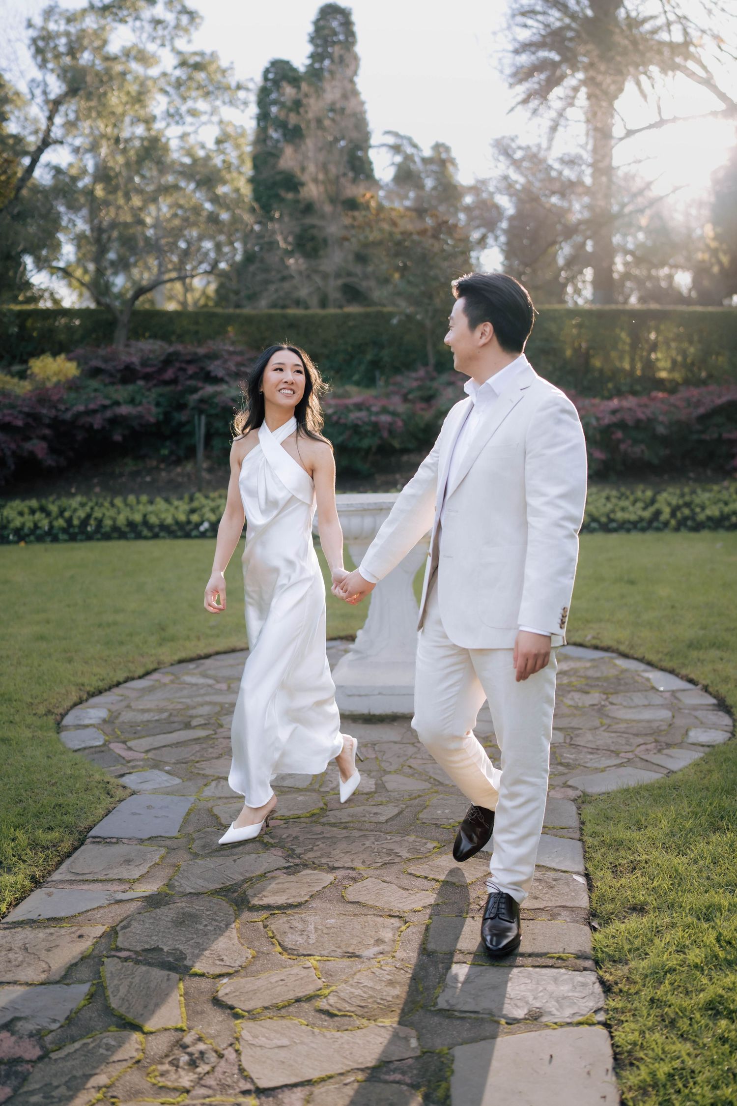 Happy couple in white wedding attire walking along a stone path in a beautiful sunlit garden.