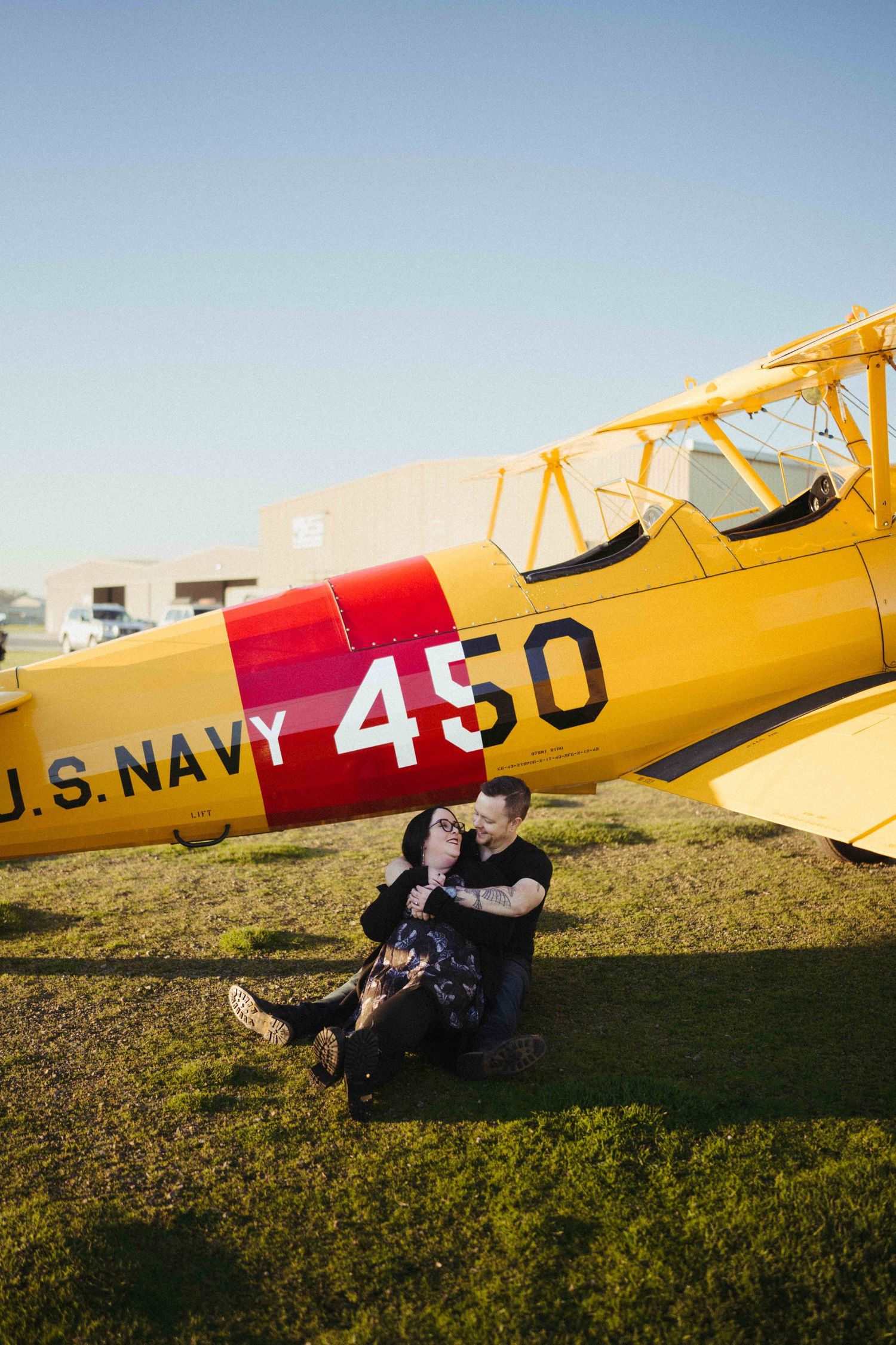Couple sitting together on grass in front of a yellow U.S. Navy biplane numbered 450 on a sunny day.