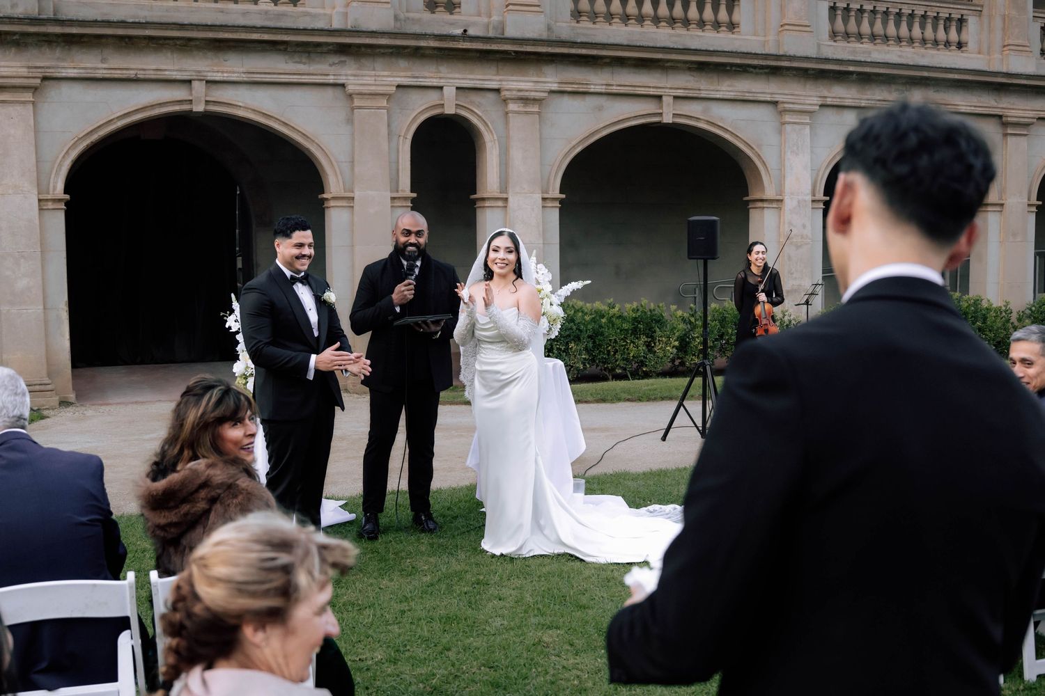 Bride and groom celebrate with officiant at outdoor wedding ceremony before arched stone building.