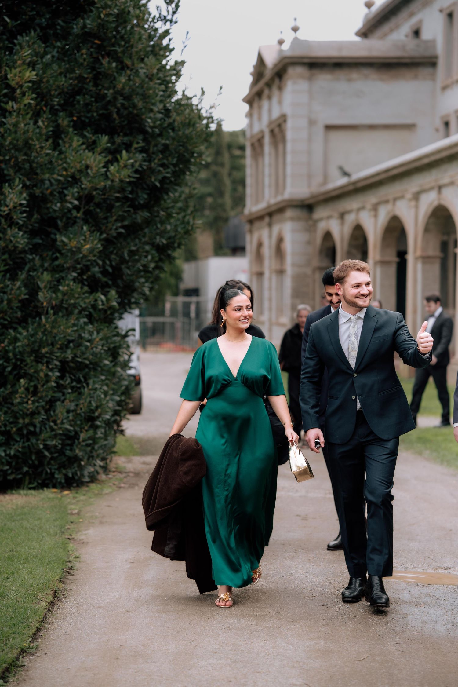 Wedding guests in elegant attire walk along garden path beside historic stone building with arched colonnade.