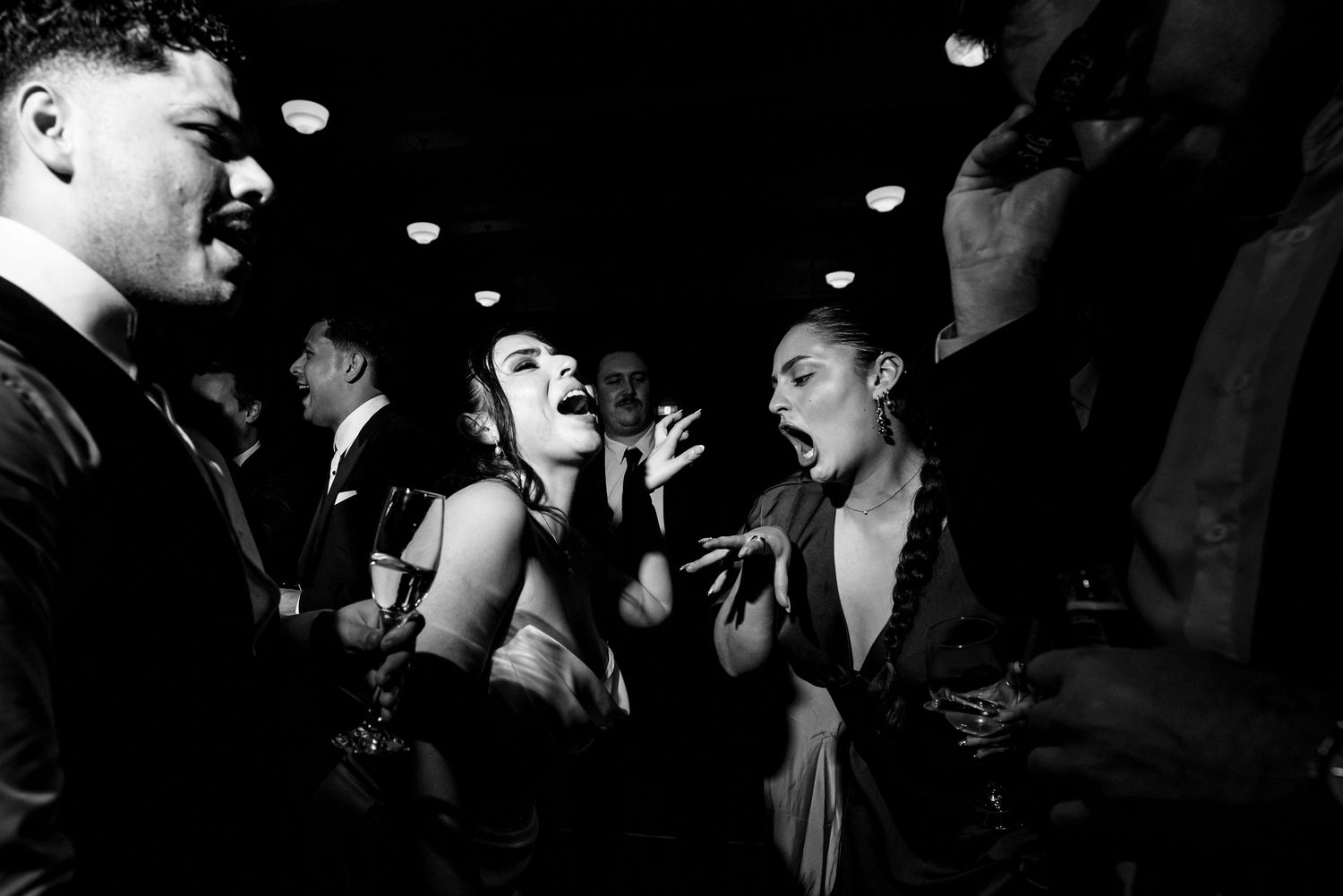 Black and white photo of women laughing and singing together on crowded wedding reception dance floor.