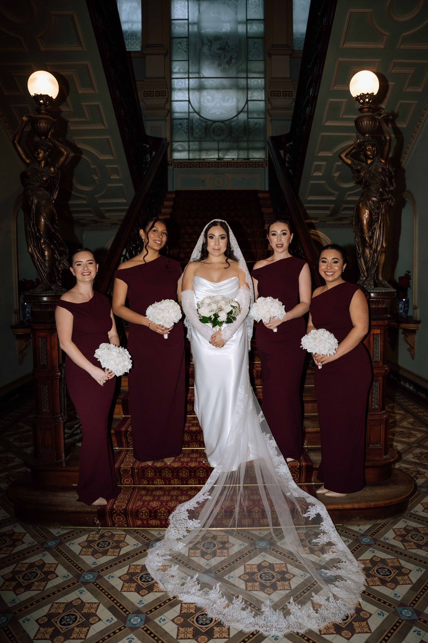Bride in white satin gown poses with four bridesmaids in burgundy dresses on ornate staircase.