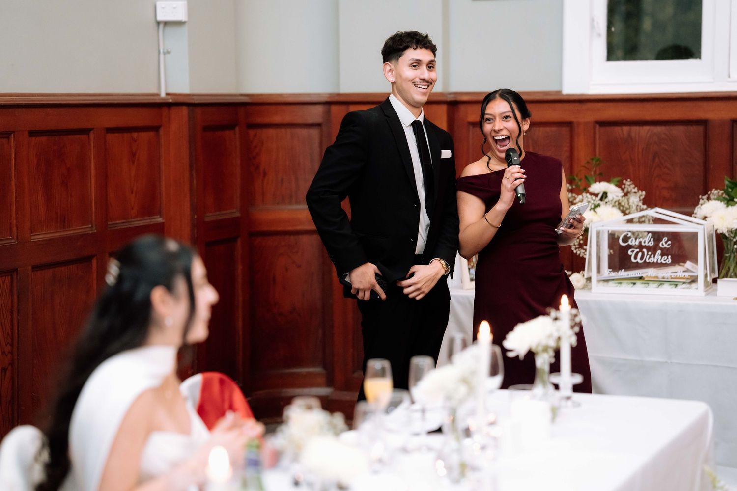 Young man in black suit and woman in burgundy dress giving speech at wedding reception.