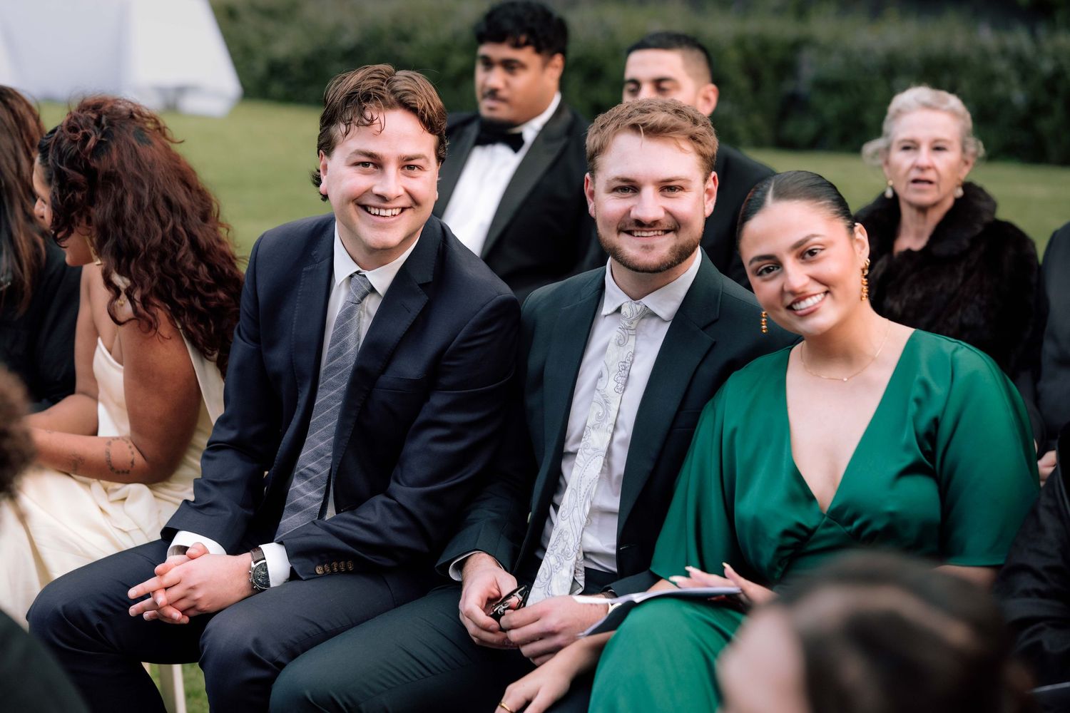 Wedding guests smiling and laughing while seated outdoors, with a woman in a green dress and two men in suits.