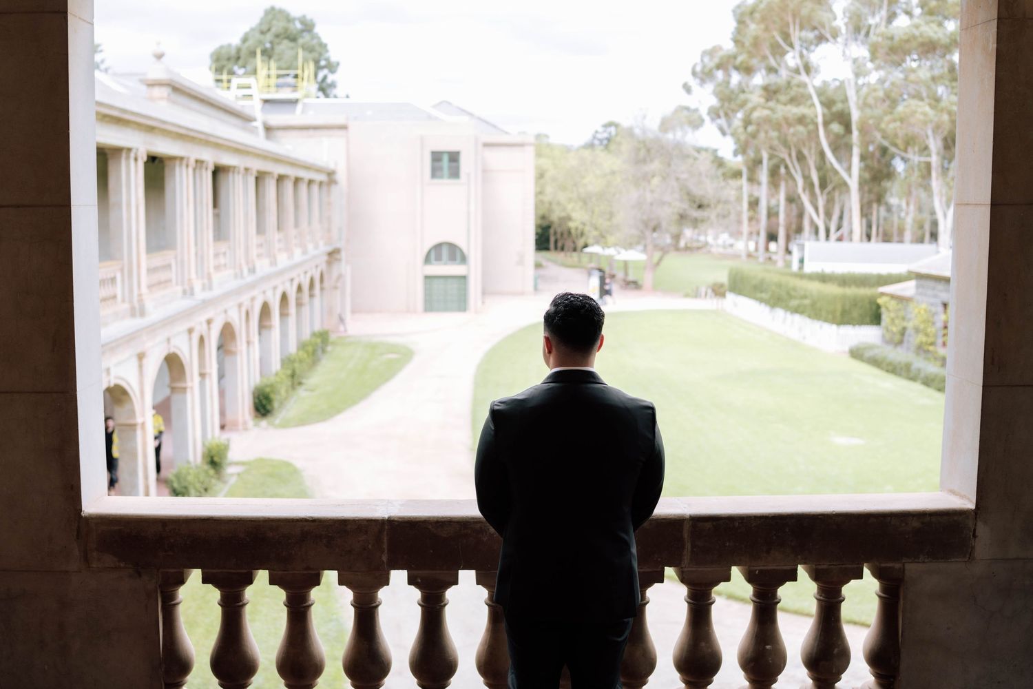 Groom in black suit stands on ornate stone balcony overlooking elegant courtyard gardens.