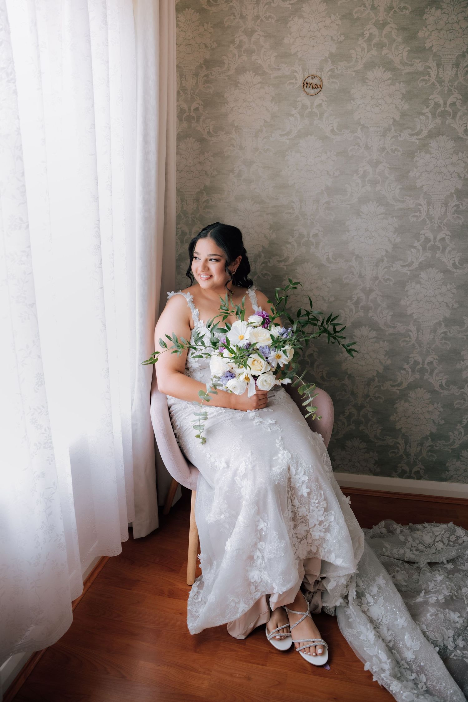 Radiant bride in lace gown seated by window holding white and lavender bouquet in elegant room.